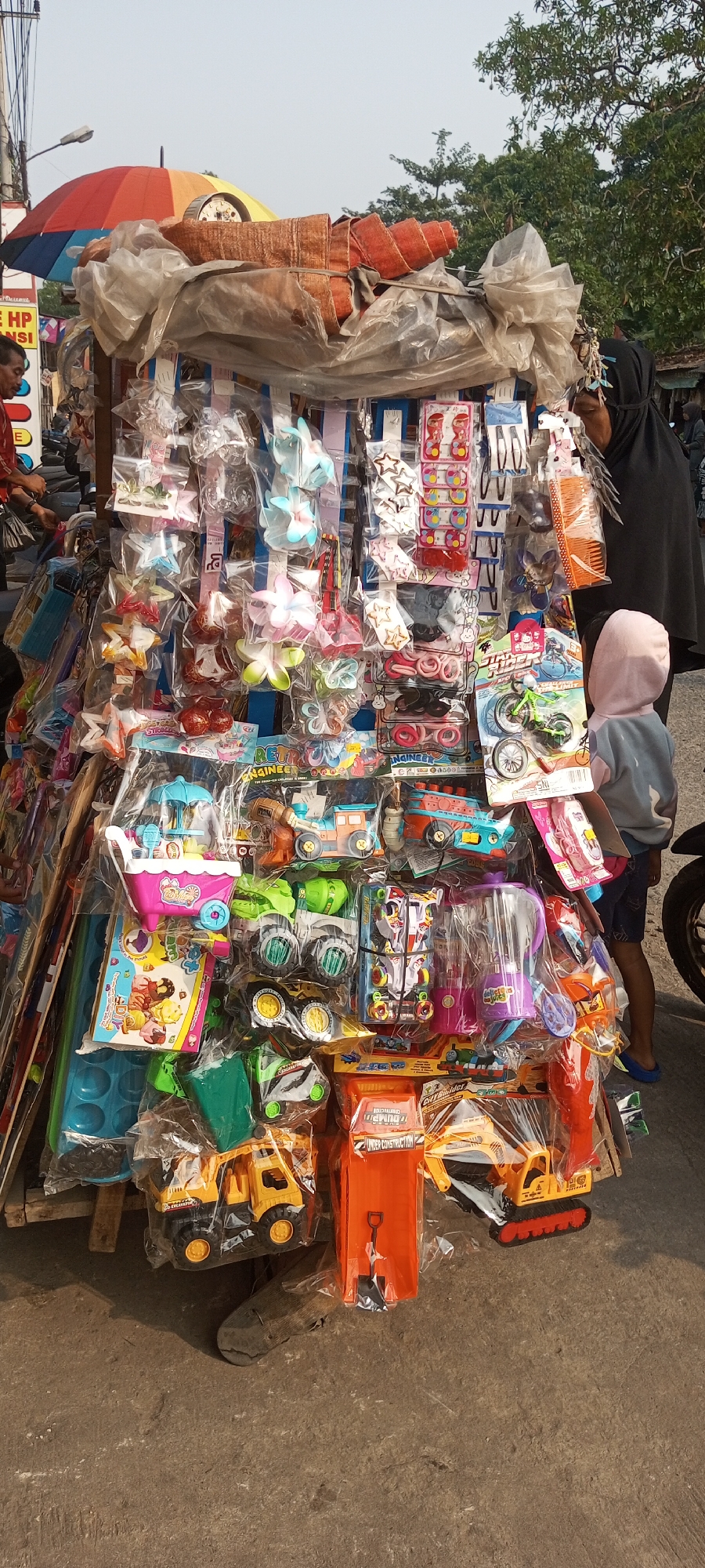 The image depicts a street vendor's cart laden with toys and accessories, situated outdoors in what appears to be a marketplace. The cart is densely packed with various items, including hair accessories like clips and headbands, small toys such as miniature bicycles, cars, and construction vehicles, as well as other miscellaneous items. The overall impression is one of a vibrant, albeit somewhat cluttered, display of merchandise.
A child wearing a light-colored hooded jacket and dark shorts stands to the right, looking towards the cart, suggesting they might be a customer. A woman wearing a black hijab and dark clothing is partially visible on the right side of the frame, also appearing to be near the cart. On the left side, a man in a red shirt is partially visible, and another person's hand is seen in the foreground. The background includes a colorful umbrella, trees, and possibly other stalls or buildings, indicative of an outdoor commercial area. The ground is paved with concrete. Based on the provided information, this scene is located in Setu, Indonesia.