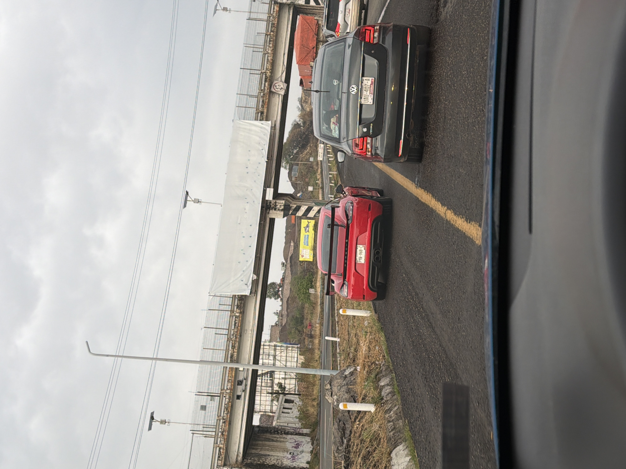 The image depicts a congested roadway in San Bernardino Chalchihuapan, Mexico, under an overcast sky. Visible are several vehicles, including a red sports car, a gray sedan, and at least one other vehicle partially visible ahead of the sedan. The vehicles are traveling on a paved road with a yellow lane divider. A bridge structure with metal framework and concrete supports runs overhead. A weathered billboard can be seen in the background near a hillside. White bollards line the side of the road nearest the sports car. The view appears to be taken from inside a vehicle, looking forward through the windshield.