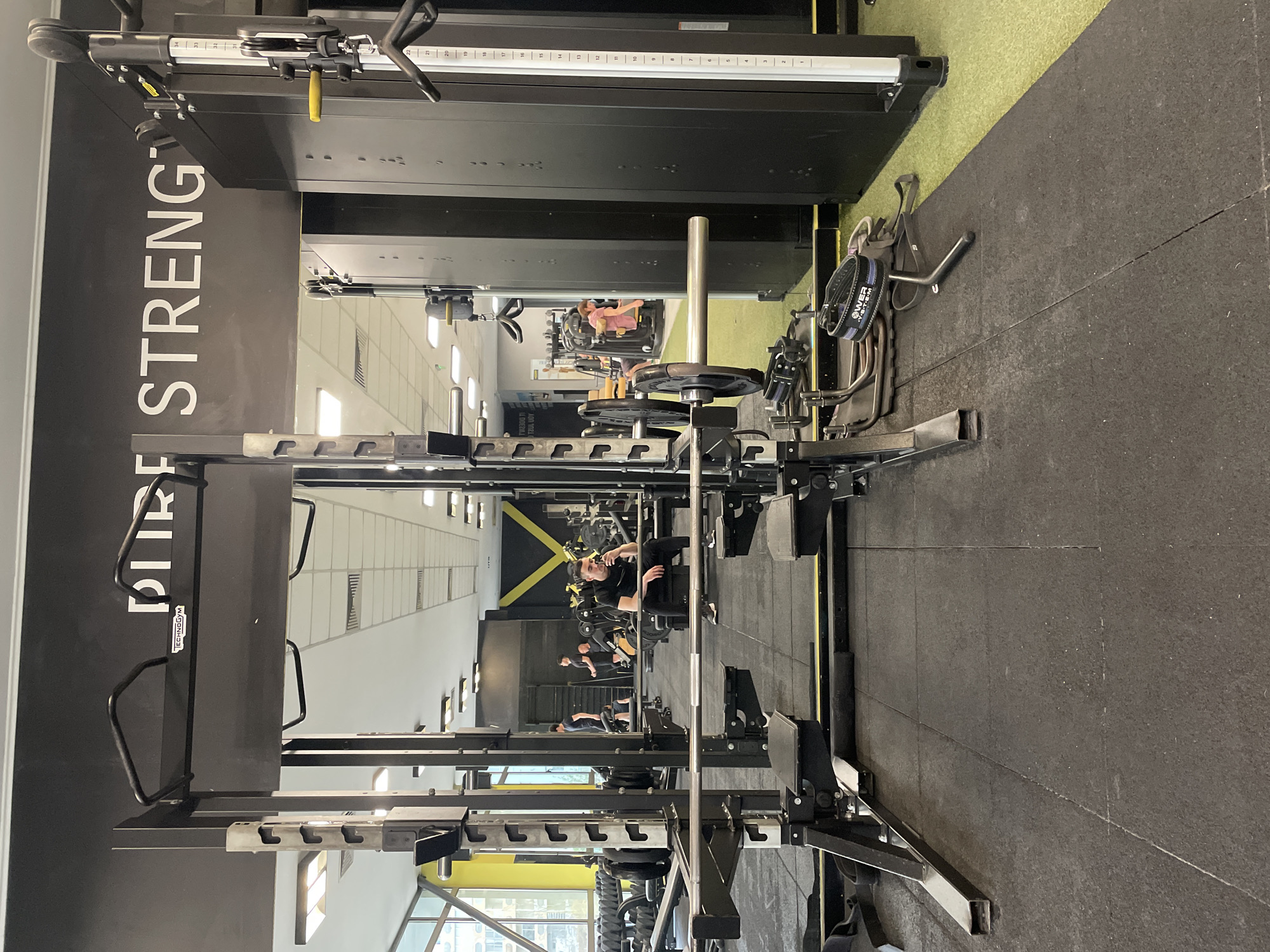 The image shows the interior of a fitness center, potentially in Plovdiv, Bulgaria, although there is no explicit confirmation. The focus is on weightlifting equipment, particularly a squat rack with a barbell loaded with weight plates. The equipment is black and silver, and the floor is covered with black rubber mats. The gym has mirrors along one wall, reflecting a row of weightlifting stations occupied by individuals engaged in workouts. The color palette includes black, grey, silver, yellow, and white. "PURE STRENGTH" is printed vertically on a large black panel. The lighting appears bright and even, suggesting a well-lit environment.