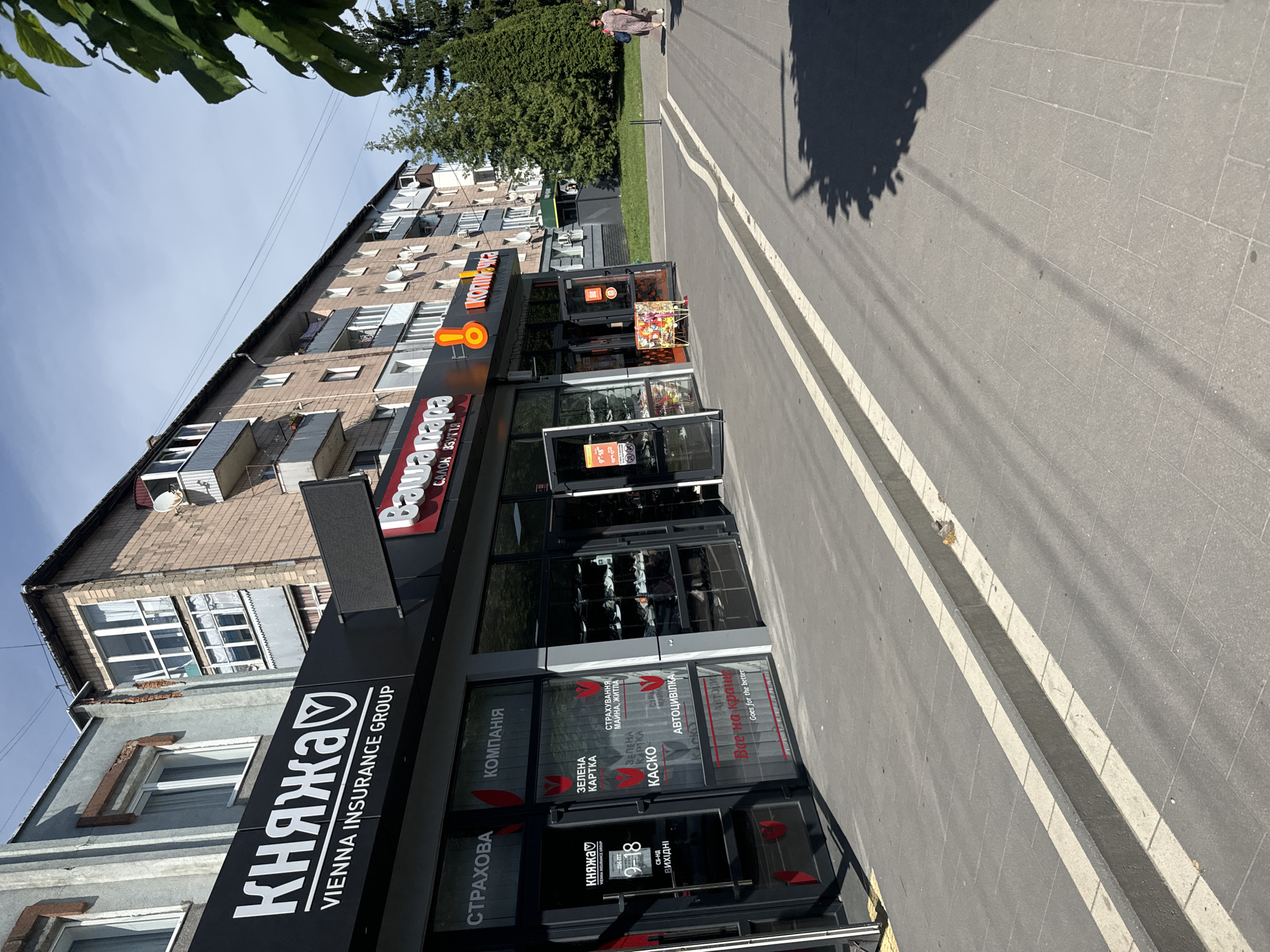 The image shows the exterior of several shops on a sunny day in the city of Sarny, Ukraine. The storefronts are modern, with large glass windows and prominent signage. One prominent sign reads "КНЯЖА VIENNA INSURANCE GROUP" in large white letters on a black background. Below this, the window displays advertisements for insurance services, including "ЗЕЛЕНА КАРТКА" (Green Card) and "КАСКО" (CASCO), with tulips depicted as a recurring motif. Another storefront has a sign that says "ВашаПА" which appears to be a shop name. Further down the street, a woman in a patterned dress and sunglasses is walking away from the camera, carrying a blue bag. The pavement is paved with grey tiles, and there is a low concrete curb with white markings. Trees and parts of residential buildings are visible in the background.