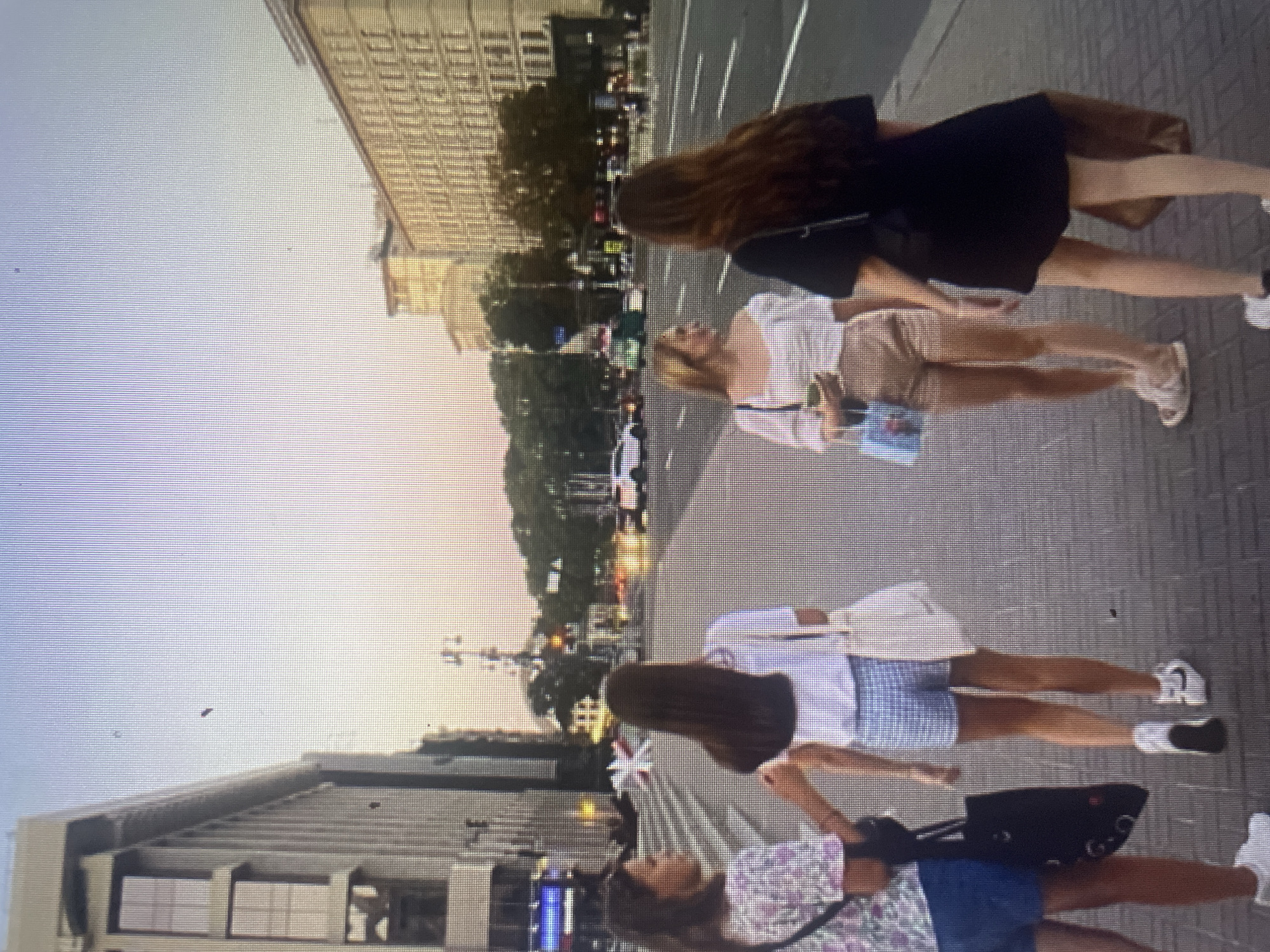 Four young women are walking on a sidewalk in a city. The woman on the far left is wearing a floral print top and blue shorts, carrying a black tote bag. Next to her, a woman in a white t-shirt and blue checkered shorts, carrying a white tote bag. The third woman, from the left, wears a white off-the-shoulder top and tan shorts, holding a drink and a blue purse. The woman on the far right is wearing a black dress and carrying a brown bag. They are walking away from the camera on a paved sidewalk adjacent to a street with traffic. Buildings line the street on both sides, and trees are visible in the background. The sky is a pale orange and blue, suggesting either sunrise or sunset. The location is identified as the city of Kołobrzeg, Poland.