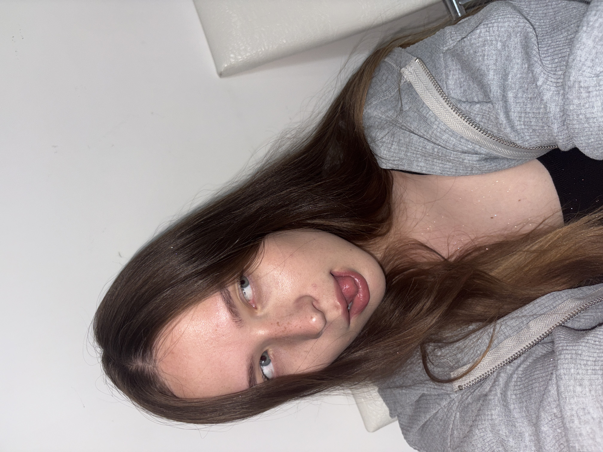 The image shows a close-up of a young woman with long, brown hair. She is wearing a grey zip-up hoodie over a black top. Her head is tilted to her left, and she is looking off to the side with her tongue sticking out slightly. Her skin has a dewy appearance, and there are some freckles visible on her nose and cheeks. Some glitter is visible on her skin and clothing. The background is a plain white wall. There is a white chair visible on the right side of the image. The image does not contain any elements that would allow for a precise location in the city of Chaikovsky, Russia.