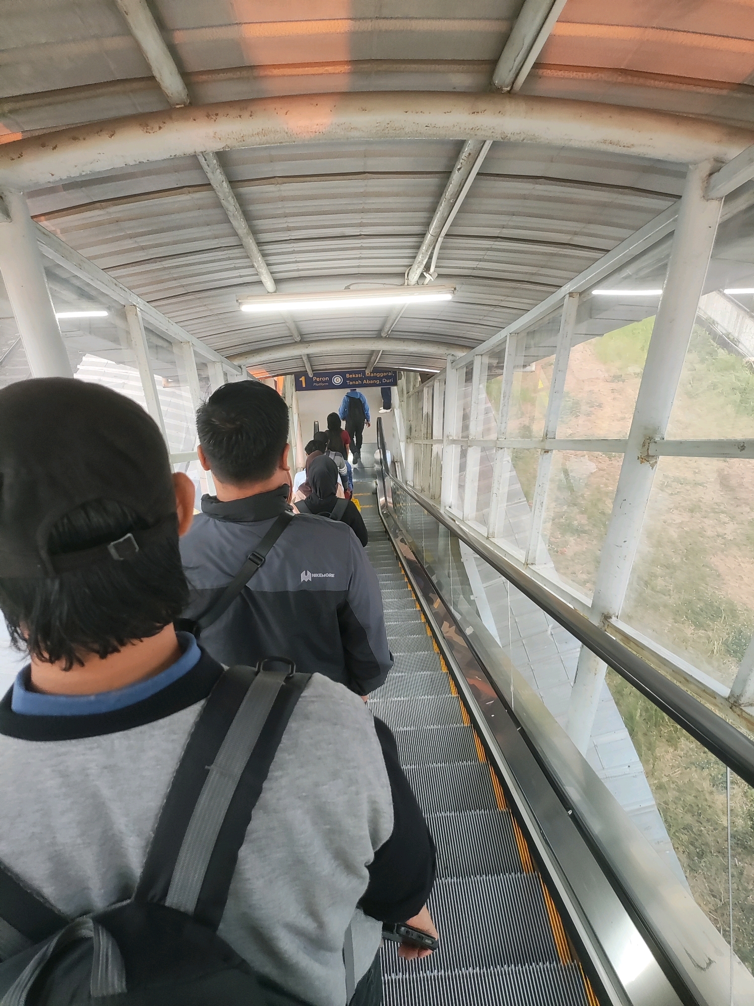 An over-the-shoulder, POV shot looks down a covered escalator at the Telagamurni train station in Cikarang Barat, Indonesia. The escalator, a moving walkway with metal treads and illuminated yellow warning strips, carries several individuals away from the camera, towards a distant platform sign. The walkway is enclosed by clear glass panels on the right, revealing an outdoor grassy area beyond. The roof structure above is made of corrugated metal supported by white painted beams and tubes. In the foreground, a person wearing a black cap and a grey sweatshirt with a backpack is visible. Further up the escalator, another person in a grey jacket with a black strap is visible, followed by other passengers. The overall scene depicts a typical transit hub environment.