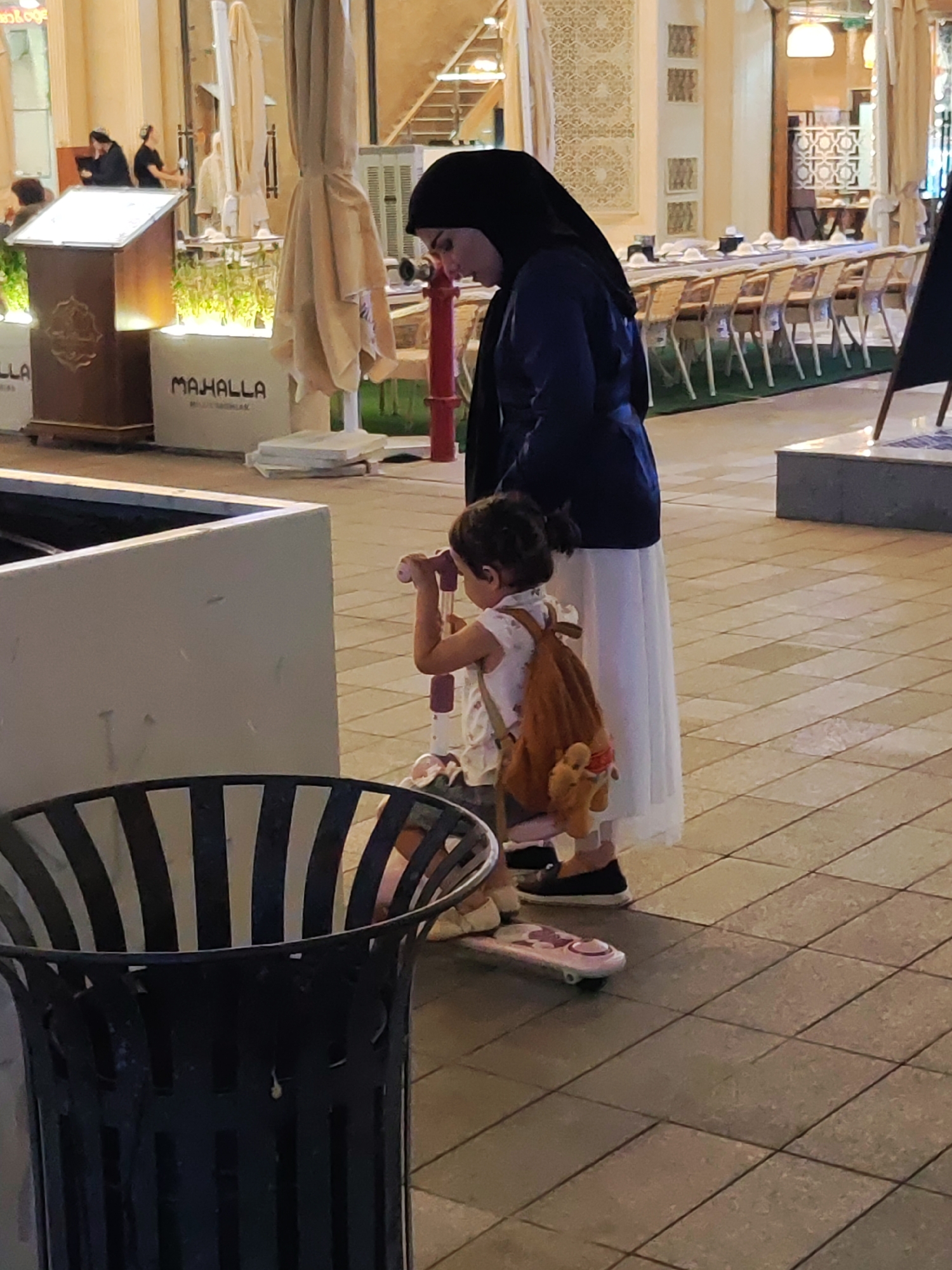 The image depicts an outdoor scene in Tashkent, Uzbekistan, likely in a pedestrian area with outdoor seating for dining establishments. In the foreground, a child is standing on a pink toy scooter. The child is wearing a light-colored dress with a pattern and has a small brown backpack with a stuffed animal attached. To the left of the child, partially visible, is a black metal trash receptacle. Behind the child and to the left, a woman is standing, wearing a dark hijab and a dark blue long coat over a white skirt. Her head is bowed, and she appears to be looking down at the child. In the background, there are outdoor tables and chairs arranged, suggesting a cafe or restaurant setting. Several large, pale umbrellas are visible, some closed. The ground is paved with square tiles. Further in the background, there are architectural elements with decorative patterns, characteristic of some buildings in Tashkent.