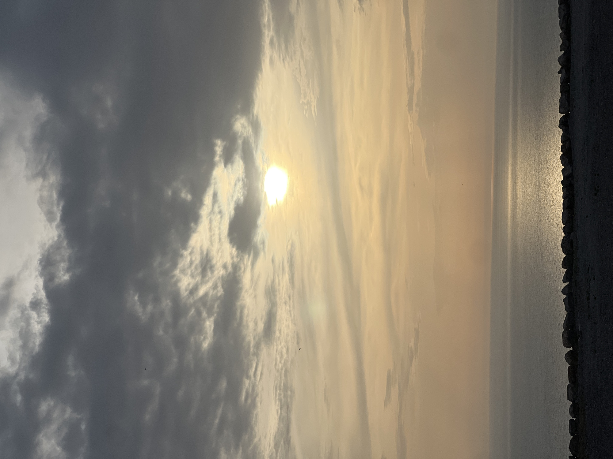The image presents a dramatic wide shot of an outdoor scene at either sunrise or sunset, featuring a vast body of water, an expansive sky, and a dark, rocky or concrete shoreline. On the right side, a rugged line of dark rocks or a man-made structure forms the edge, separating the water from the land or a path, with some surfaces subtly reflecting the sun's light.
The sky is largely dominated by clouds. On the left, heavy, dark grey clouds create a dense, somewhat foreboding atmosphere, while towards the center and right, the clouds appear lighter and more wispy. The sun, a bright and intense white-yellow orb, is prominently featured, partially obscured by these lighter clouds. Its low position in the sky is characteristic of dawn or dusk.
The sun casts a brilliant, shimmering path of light across the calm surface of the water, extending from the horizon towards the viewer. The rest of the water appears tranquil and a muted grey-blue, reflecting the sky above. Several small, dark specks, likely birds, are visible against the cloudy sky. The overall impression is one of serene power and natural beauty, highlighting the contrast between the dark, heavy clouds and the intense, warm light of the sun. There is no visible text in the image. The location context provided is Kovačko Polje, Montenegro, implying this is a coastal scene.