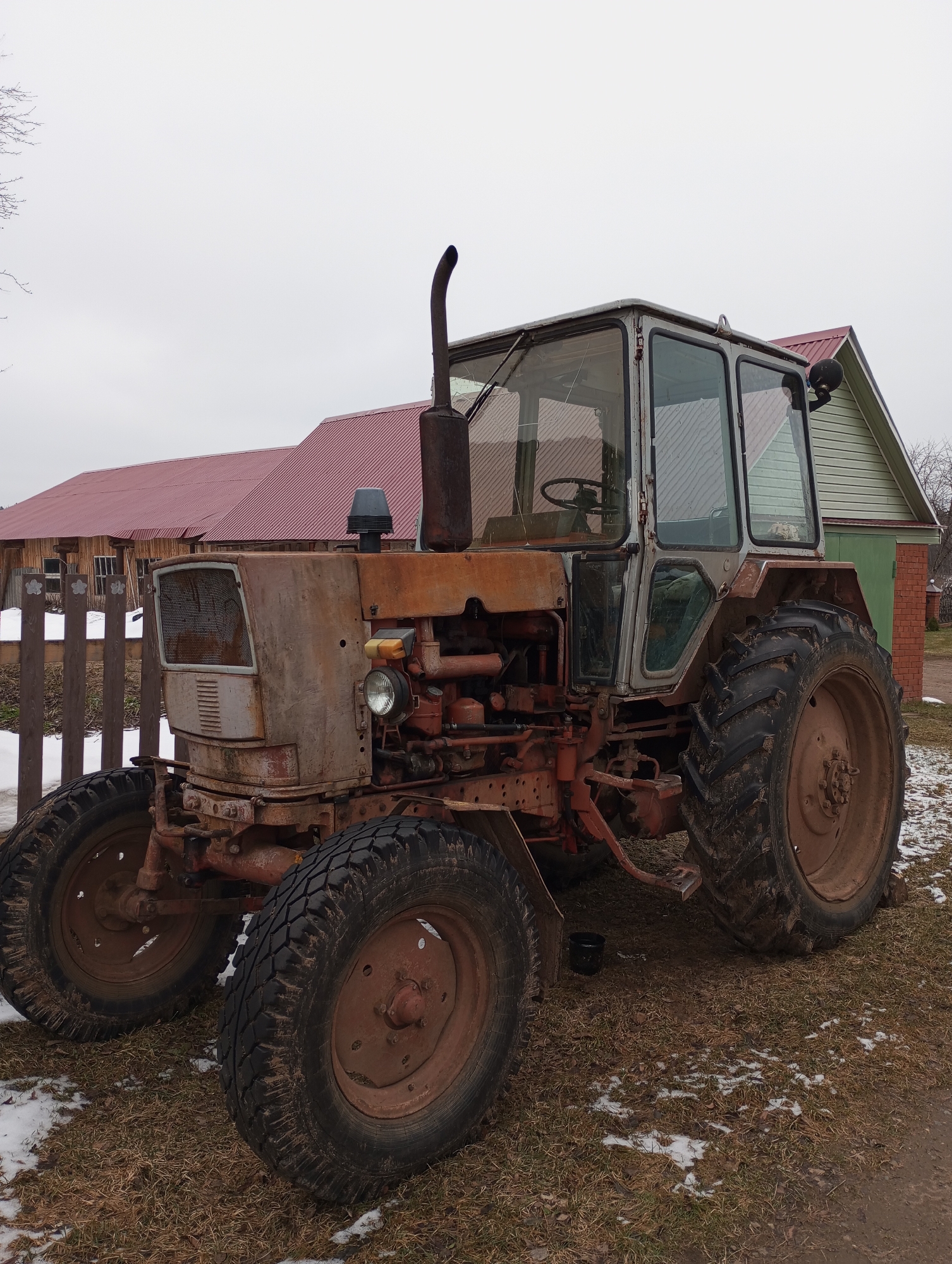 The image depicts a weathered, vintage tractor parked in what appears to be a rural or semi-rural setting. The tractor is predominantly rust-colored with a grey cab, featuring large, mud-caked tires. The tractor's condition suggests it is old and possibly disused. Behind the tractor, there is a wooden fence and two structures: one with a red metal roof and light-colored wooden siding, and another with a green facade and a red brick section visible. The ground around the tractor is covered in dry grass and patches of snow, indicating it is likely winter or early spring. The scene overall suggests a farm or agricultural environment. While the location cannot be definitively confirmed as Буренка, Russia, the architecture and the rural scene are consistent with images of Russian villages.