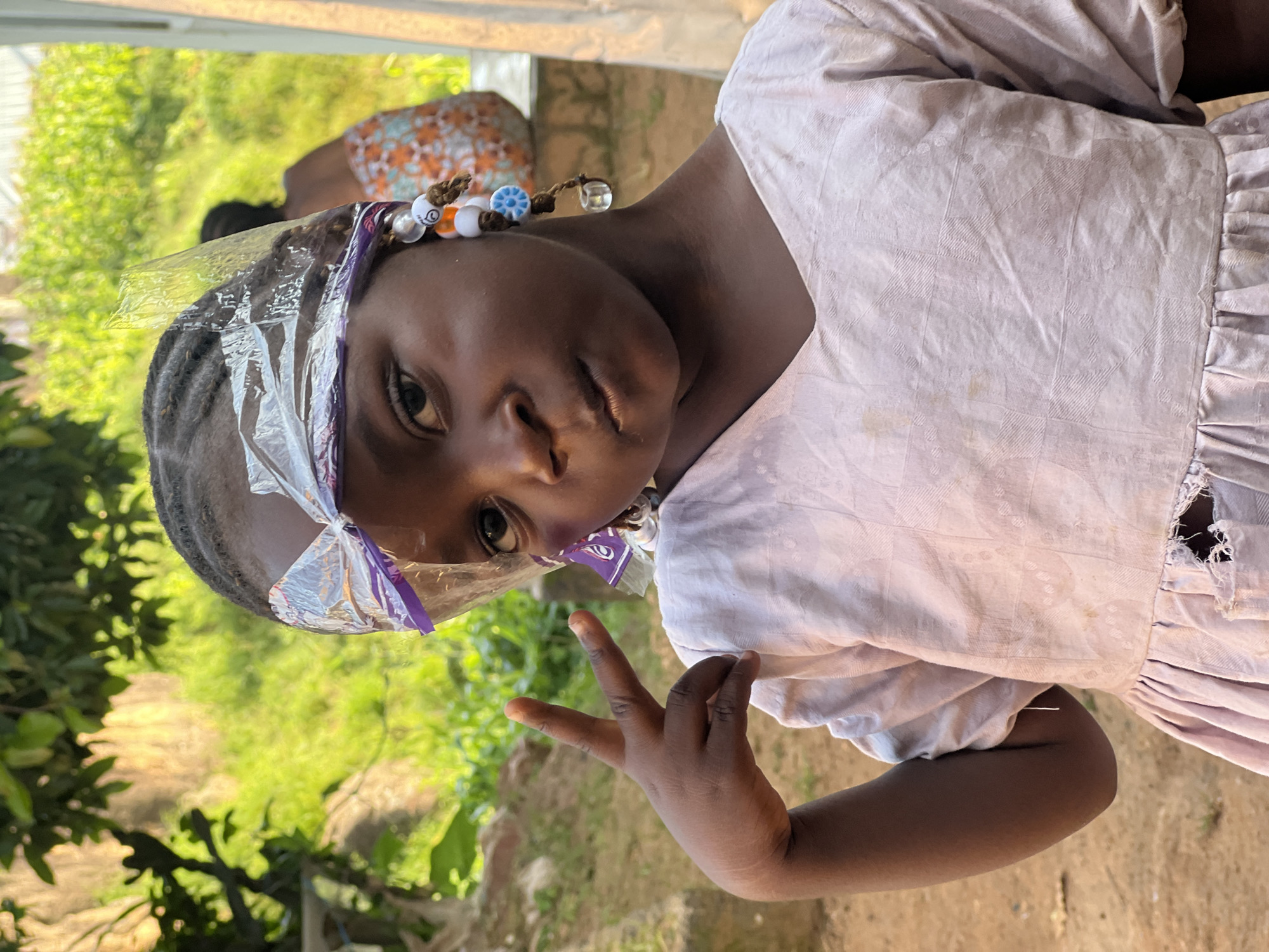 A young girl, appearing to be of African descent, is pictured in a medium close-up shot outdoors. She has dark skin and her hair is braided and wrapped with a metallic-looking material, possibly foil, secured with a purple ribbon. She is wearing a light-colored dress with a V-neckline and short, puffed sleeves. The dress appears to be made of a textured fabric and has some damage, with a tear visible near the hemline. The girl is holding up her left hand, displaying the peace sign with her index and middle fingers. She has beads in her hair, hanging from her ear area, including white, orange, and blue beads, with one clear bead at the end.
In the background, out of focus, a person in a colorful, patterned wrapper is visible, sitting or leaning against a structure. Lush green vegetation and trees are also present, suggesting a natural or semi-rural environment. The ground in the foreground is a reddish-brown color, consistent with soil. The lighting suggests it is daytime, with sunlight illuminating the scene.
While the image itself does not provide explicit clues to its location within Jos, Nigeria, the overall context of the girl's appearance, the clothing styles, and the vegetation are consistent with regions in Nigeria.