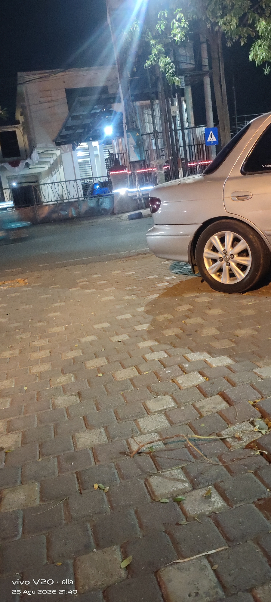 This image captures a street scene at night, likely in Tulungagung, Indonesia, as indicated by the date stamp "25 Agu 2026 21.40". The foreground is dominated by a paved walkway made of interlocking rectangular bricks, some of which appear damp or stained. Scattered on the walkway are a few fallen leaves and twigs, suggesting an outdoor environment.
In the mid-ground, a silver sedan is parked, its rear section clearly visible. The car has a clean design, with a prominent alloy wheel featuring a multi-spoke pattern. The vehicle is positioned on the street adjacent to the walkway.
Behind the car, a building facade is visible, illuminated by artificial lights. The building features a modern architectural style with clean lines and what appears to be glass panels or windows. A fence with decorative metalwork separates the building from the street.
Further in the background, there are more structures and what looks like streetlights or spotlights casting bright beams of light. A blue traffic sign with a white pedestrian symbol is also visible, mounted on a pole, indicating a pedestrian crossing or zone. Trees with green foliage are present, their leaves catching the light. The overall atmosphere is dimly lit, typical of a nighttime urban setting. No people are directly depicted in the image, but the presence of the pedestrian sign implies human activity in the vicinity.