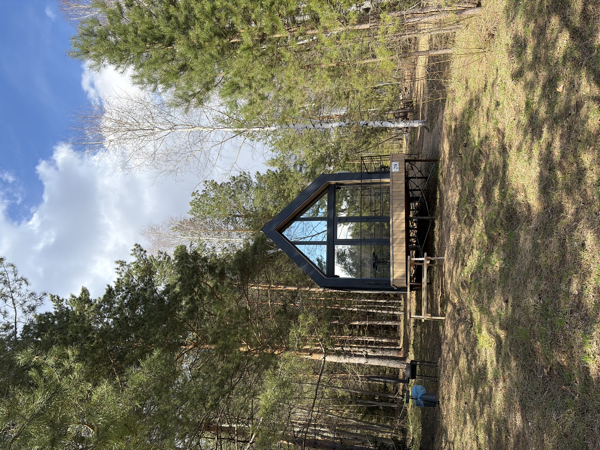 The image depicts a modern A-frame cabin situated within a forest clearing in Мишкино, Russia. The cabin, constructed with a dark grey frame and light wooden siding, features a distinctive triangular shape with large, expansive glass windows on its front facade, reflecting the blue sky and surrounding pine trees. It stands on an elevated wooden deck, beneath which additional wooden supports are visible. A black metal railing lines the deck, where a dark metal outdoor table and chair are visible. A white plaque bearing the number "20" is affixed to the cabin's exterior near the deck.
The surrounding environment is a dense forest, dominated by tall coniferous trees, primarily pines with their characteristic green needles. Scattered among them are several deciduous trees, notably birches, identifiable by their white bark and bare branches, suggesting late winter or early spring. The ground is covered with dry grass and patches of earth, with numerous tree shadows cast across it from the bright sunlight. Near the base of the cabin, a blue plastic bag, possibly a garbage bag, rests on a black stand.
The scene appears to be captured during the daytime, under a bright sky with scattered white cumulus clouds. There are no people visible, and no specific activity is taking place, presenting a tranquil and still moment of a modern dwelling integrated into a natural, forested landscape.
