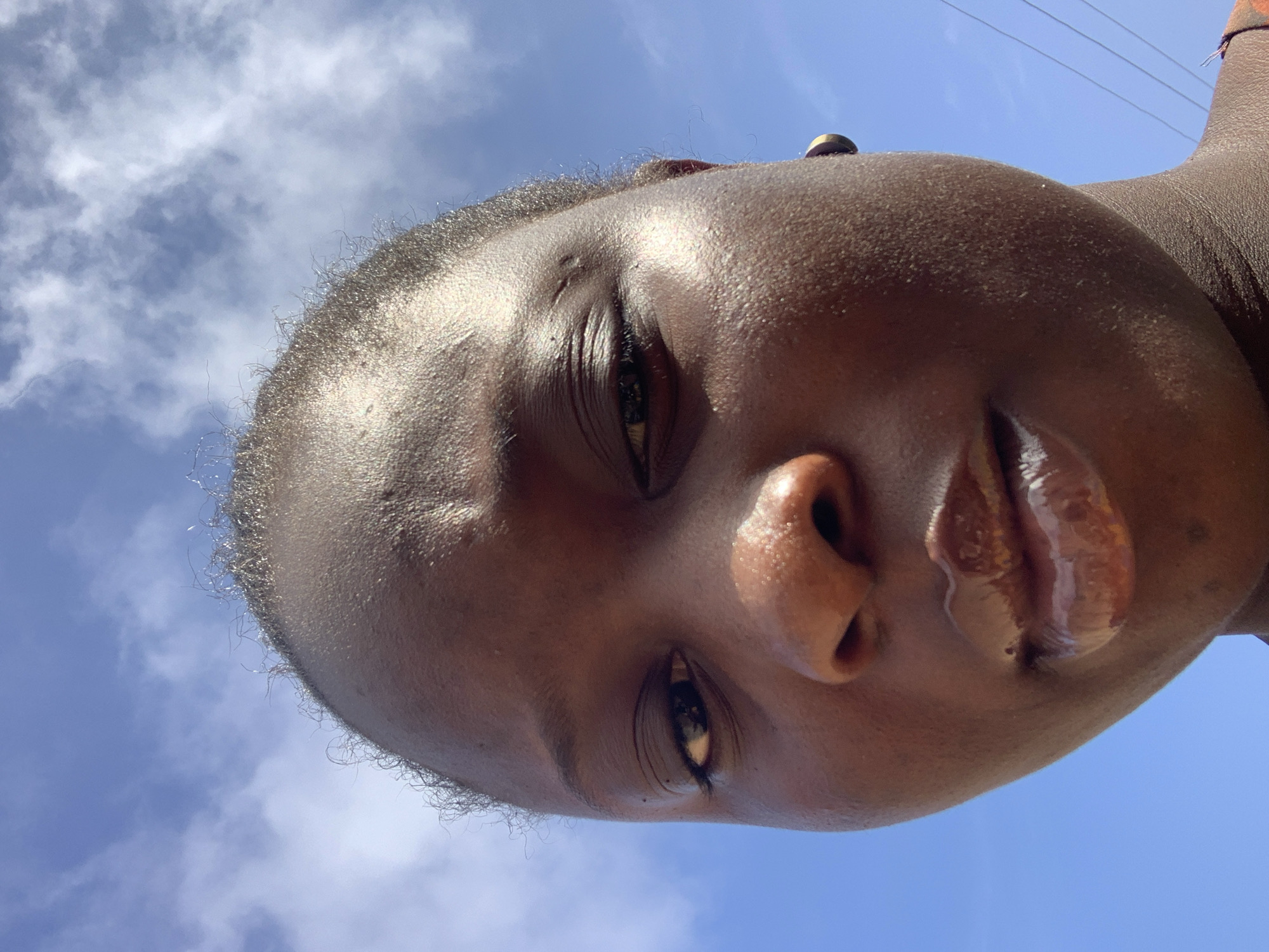 A close-up, low-angle selfie shows the face of a person against a bright blue sky with scattered white clouds. The person's face is dark-skinned and angled slightly down towards the camera. Their forehead is visible at the top of the frame, with wisps of dark hair at the hairline. The eyes are dark and appear to be looking directly at the camera. The nose is prominent, and the lips are full and glossy, reflecting the sunlight. A small stud earring is visible in the person's right ear. The skin appears slightly shiny, suggesting it may be warm or humid. The background is predominantly sky, with hints of what might be power lines or distant structures on the far right edge of the image. The overall impression is one of an outdoor shot taken on a sunny day, with the camera positioned below eye level, looking upwards at the subject.