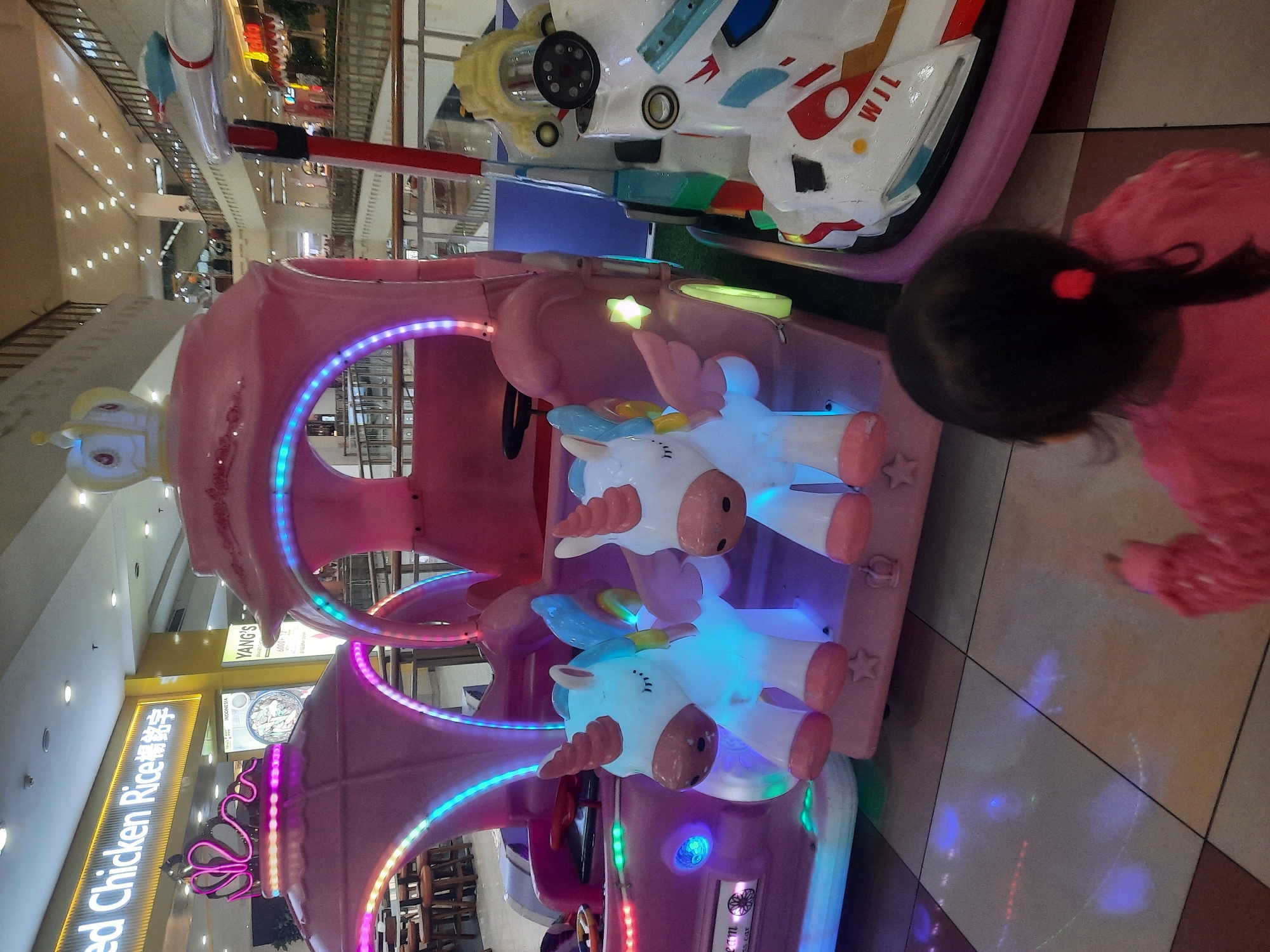 The image depicts a child from behind, wearing a pink sweater with a red hair tie, looking at a row of brightly lit, pink unicorn-themed amusement park rides. The rides are designed to resemble carriages and have LED lights in various colors. In the background, there is a mall interior with multiple levels, railings, and storefronts. A prominent sign in yellow states "Red Chicken Rice" in English and Chinese characters, indicating the location is likely a food establishment within the mall. Another sign reads "YANG'S" and displays food items. The floor is tiled in a checkered pattern of light and dark squares, with reflections of the lights from the rides visible on the floor. The scene is set in a commercial environment, likely a shopping mall in North Jakarta, Indonesia, given the presence of Indonesian text on signs.