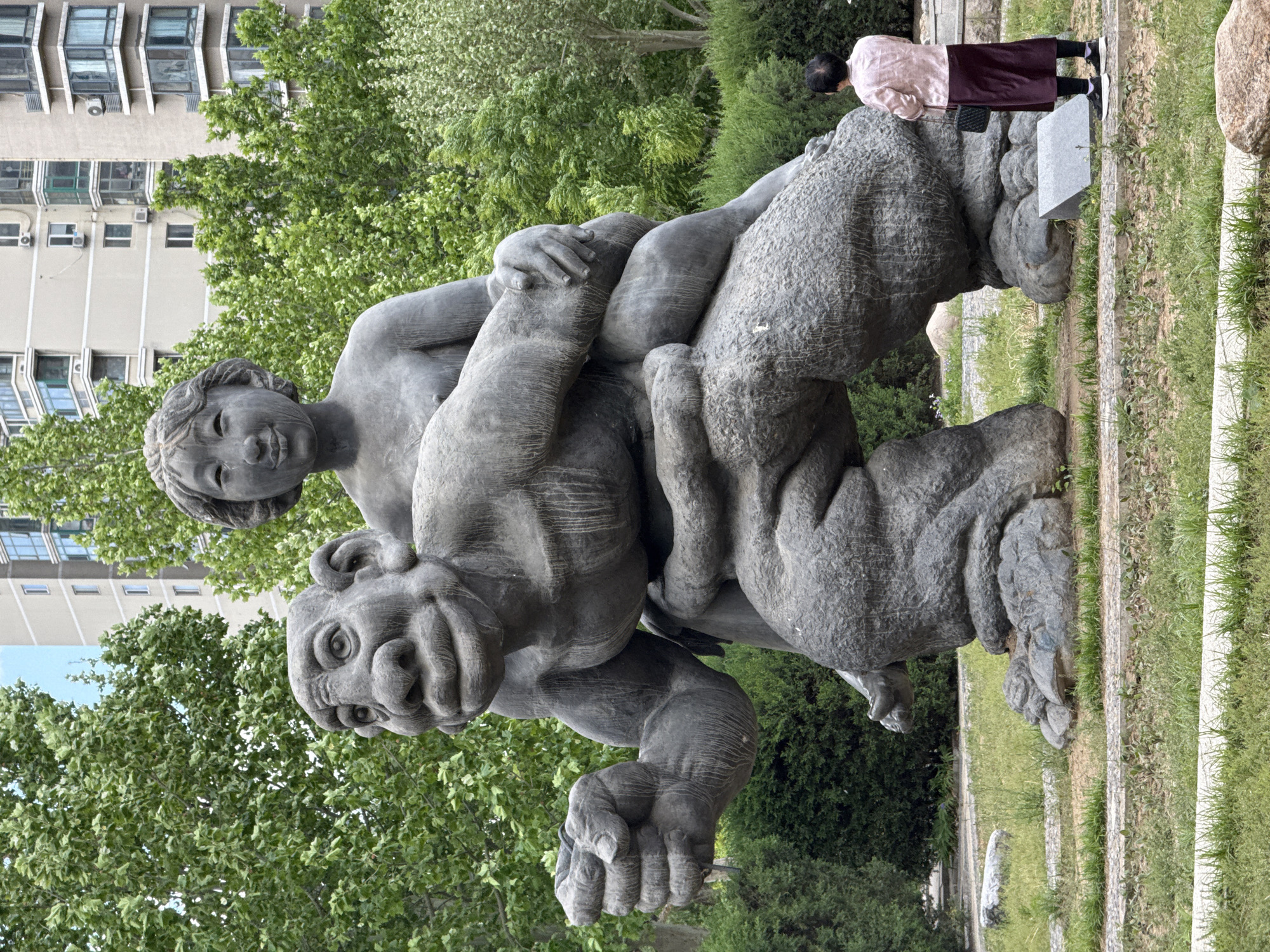 Here's a detailed description of the image:
**Overall Impression:** The image shows a large stone sculpture of a man carrying another person on his shoulders. The sculpture is placed outdoors, in what appears to be a park or garden setting. A person stands on the ground next to the sculpture. Tall buildings and lush greenery can be seen in the background. The image is oriented vertically.
**Sculpture Details:** The sculpture depicts two figures. The lower figure is larger, more muscular, and has a somewhat grotesque facial expression. The upper figure is smaller, younger, and seems to be embracing the larger figure. The style is figurative but with some level of abstraction. The material appears to be a rough-hewn stone, possibly granite or similar.
**People:** An individual is standing near the sculpture. The person is wearing a pink or light-colored shirt and a dark skirt. The person is standing on the ground, on the edge of a lawn.
**Environment:** The sculpture is placed in an outdoor setting, possibly a park or garden. There's grass and other greenery around the base of the sculpture. In the background, there are tall apartment buildings and trees.
**Location:** Based on the image and available information, it is likely the location is in a city in Changping District, China. The buildings in the background suggest a developed urban area.