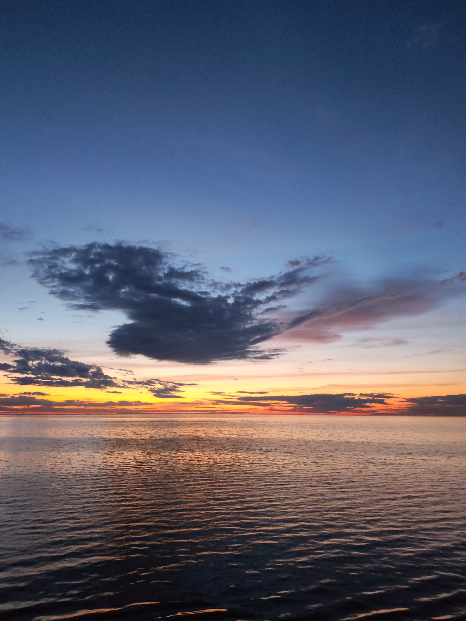 The image depicts a serene seascape at dusk or dawn. The sky transitions from a deep blue at the top to a warm orange and yellow horizon. Several cloud formations are visible, with a prominent dark cloud cluster towards the left side of the image and lighter, streaky clouds towards the right. The sea reflects the colors of the sky, with gentle ripples creating a textured surface. There are no discernible people or objects other than the sky and the sea. The location is unknown.