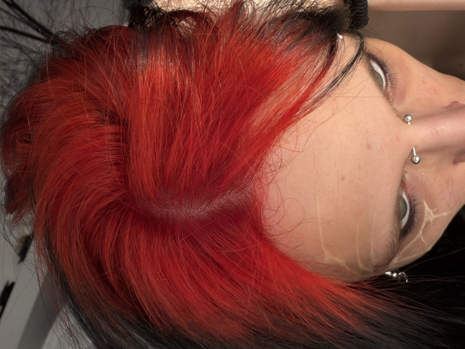 The image displays a high-angle, close-up perspective of an individual's head and upper facial features. The hair presents a prominent, vibrant red coloration at the top and crown, transitioning to darker, likely black, hair along the sides and visible underlayers. A central part is discernable in the scalp. The forehead exhibits several faint, pale, linear marks resembling superficial scarring or decorative application. The individual's eyes are visible, looking upward towards the camera, with dark cosmetic eyeliner applied. A silver barbell piercing is present on the left eyebrow. A spherical silver piercing element is visible on the bridge of the nose, consistent with a septum piercing. A small, silver, dangling earring is visible near the individual's left temple area. The background is out of focus, appearing as an indistinct, light-toned area, indicative of an indoor environment. The specific location within the city of Plovdiv, Bulgaria, cannot be determined from the visual information provided in the image.