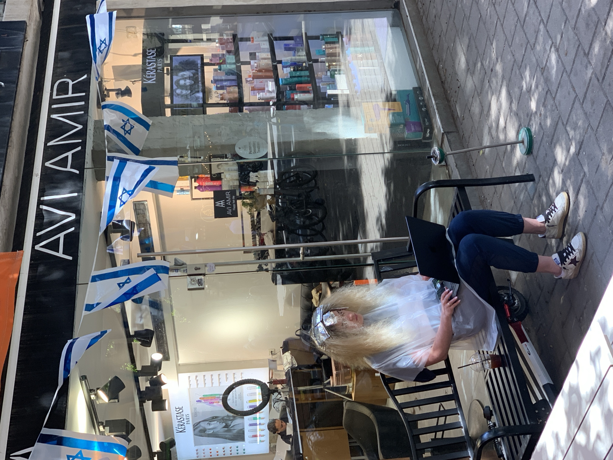 A bright outdoor scene captures a person getting a hair treatment while using a laptop, in front of a hair salon in Tel-Aviv, Israel. The person, with long, light blonde curly hair, is wearing a white plastic smock, dark pants, and white sneakers with a red star design. They are seated on a black metal bench, focused on a black laptop. An electric scooter is parked beneath the bench, and a plastic cup with a straw containing a drink rests on the ground nearby.
The storefront is adorned with numerous small Israeli flags strung across its facade. Large glass windows reflect the sunny surroundings and reveal the interior of what appears to be a hair salon or beauty supply store. Inside, shelves are stocked with various hair care products, prominently featuring "KÉRASTASE PARIS" branding. Other visible texts include "AVI AMIR" on the main sign and a smaller sign inside, which also reads "Happy hair day" and "Permanent Smoothing Treatment." A display showing a woman's face and a ring light are also visible through the glass.
The ground is a paved sidewalk, showing distinct shadows, indicating bright daylight. A tall, slender ashtray stand with green accents is positioned on the sidewalk to the right. The overall impression is a bustling urban setting, where daily activities like hair care and work converge.