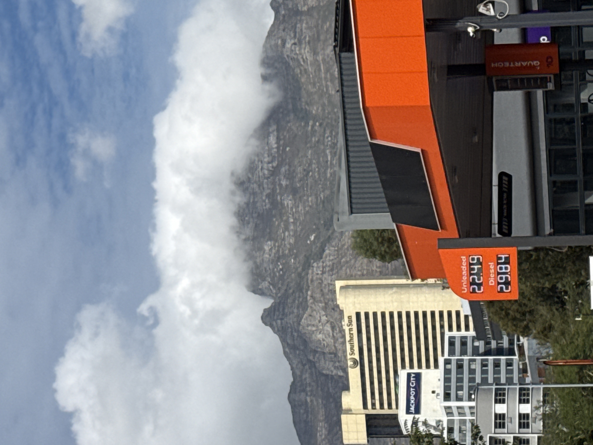 The image depicts a view in Cape Town, South Africa. Table Mountain dominates the background, partially obscured by clouds. A petrol station sign displays fuel prices: Unleaded at 22.49 and Diesel at 29.84. Adjacent to the petrol station are buildings, including "Southern Sun" and "Jackpot City." The sky is partially cloudy.