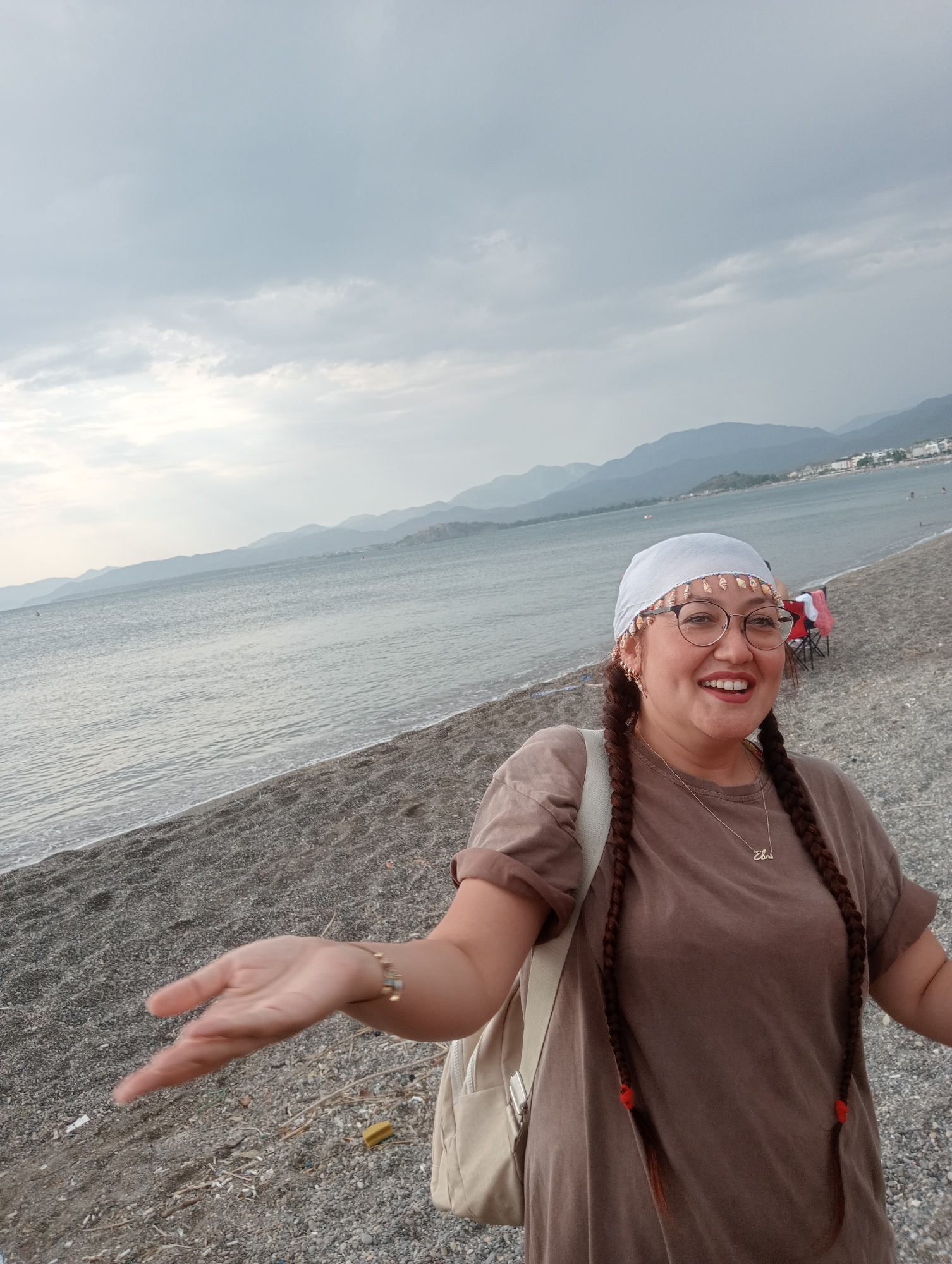 A medium shot shows a woman with braided hair, glasses, and a white headscarf standing on a beach in Fethiye, Turkey. She is smiling and has her left arm outstretched. The beach is covered in pebbles, and the sea is visible in the background, with mountains in the distance. The sky is overcast with clouds.