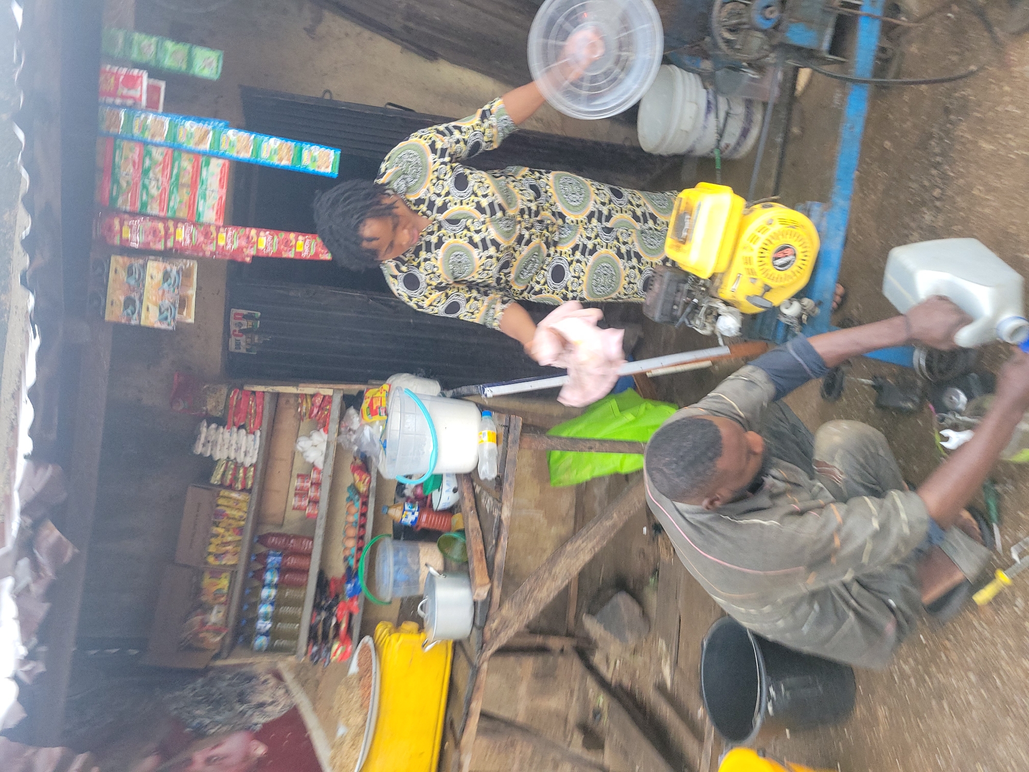 The image depicts an outdoor scene, likely at a market stall, with a woman and a man engaged in activities. The stall itself appears rustic, constructed from wood and covered with a corrugated metal roof. Shelves are stocked with various goods, including bottled beverages, packaged food items, and what appear to be cartons of noodles or similar products.
In the foreground, a man wearing a grey work jumpsuit is crouched down, appearing to be working on a yellow engine or generator. He is holding a white plastic container with a blue cap in his left hand and a white object that might be a rag or a piece of cloth in his right hand, near the engine. Tools, including a wrench, are visible on the ground around him.
A woman, dressed in a patterned dress, stands behind the man and the engine. She is holding a light pink cloth in her left hand and a clear plastic lid or plate in her right hand. Her attention is focused downwards, towards the engine area.
The environment surrounding the stall is unpaved and appears to be muddy or dusty. Several buckets are scattered around, including a black bucket and a yellow bucket. A large, flat yellow container with granular material is positioned on a wooden surface to the left of the scene. In the background, a doorway with a dark metal gate is visible. The overall impression is one of a working market environment in a somewhat utilitarian setting.