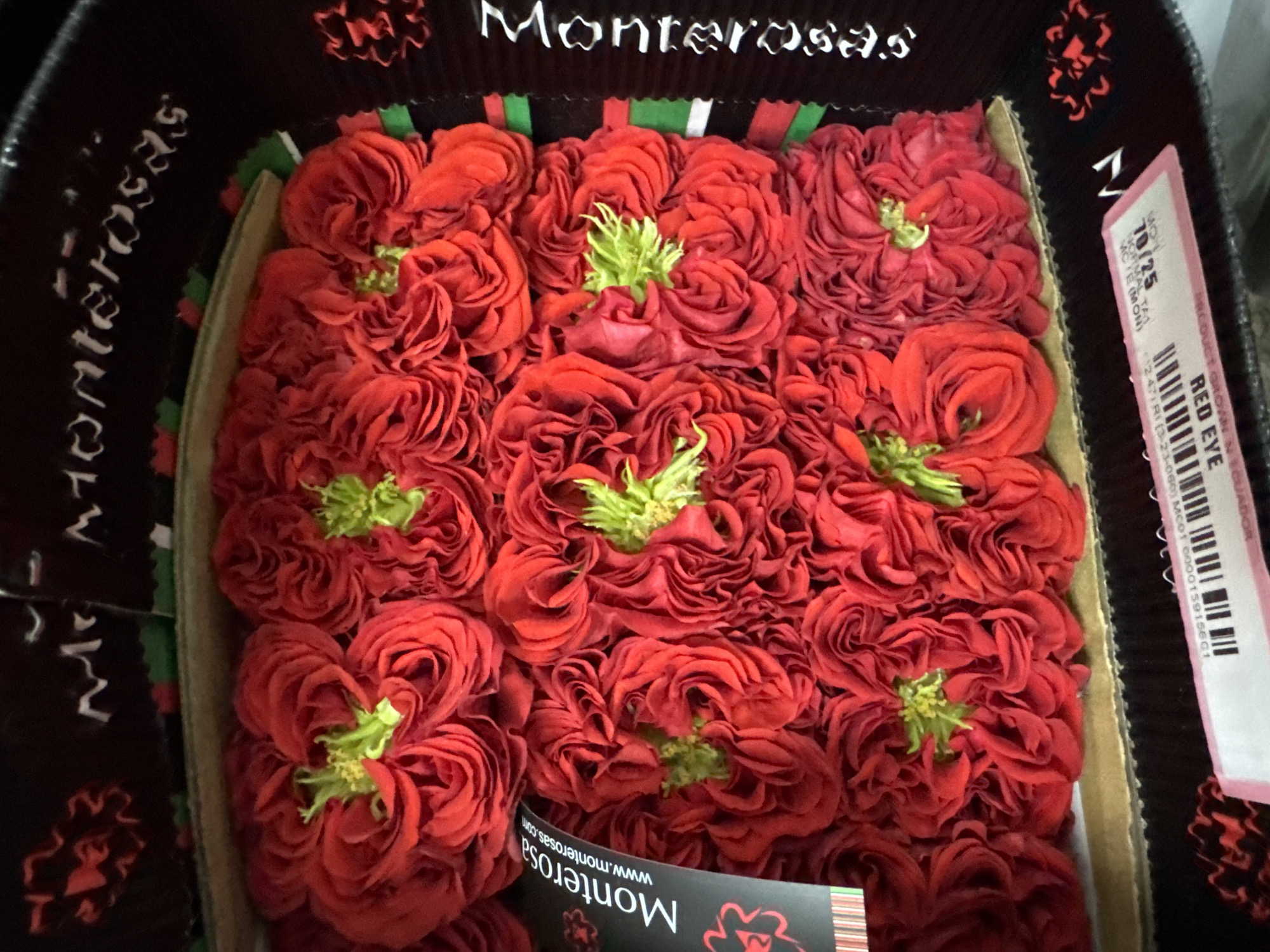 The image shows a box of red roses. The roses are tightly packed and appear to be fresh. The box is black with red and green accents. There is a label on the side of the box that says "Monterosa" and "www.monterosas.com". There is also a pink tag with a barcode and the words "RED EYE". The image does not provide any information about the location in Izhevsk, Russia.