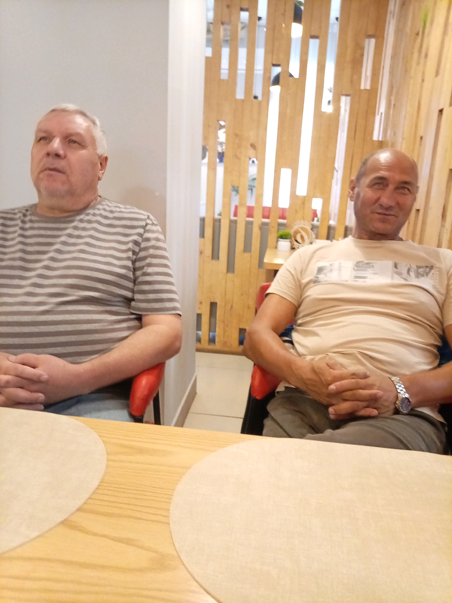 Two men are seated at a table in what appears to be a restaurant or cafe. The man on the left is older, with grey hair and a striped t-shirt. He is looking upwards and to the left. The man on the right is younger, bald, and wearing a light-colored t-shirt and a watch on his left wrist. He is looking towards the camera. Both men have their hands clasped in front of them. They are seated in red chairs. In the background, there is a decorative wooden wall with vertical slats and some lighting. The table in front of them is made of light-colored wood and has two round, light-colored placemats. The scene appears to be indoors.