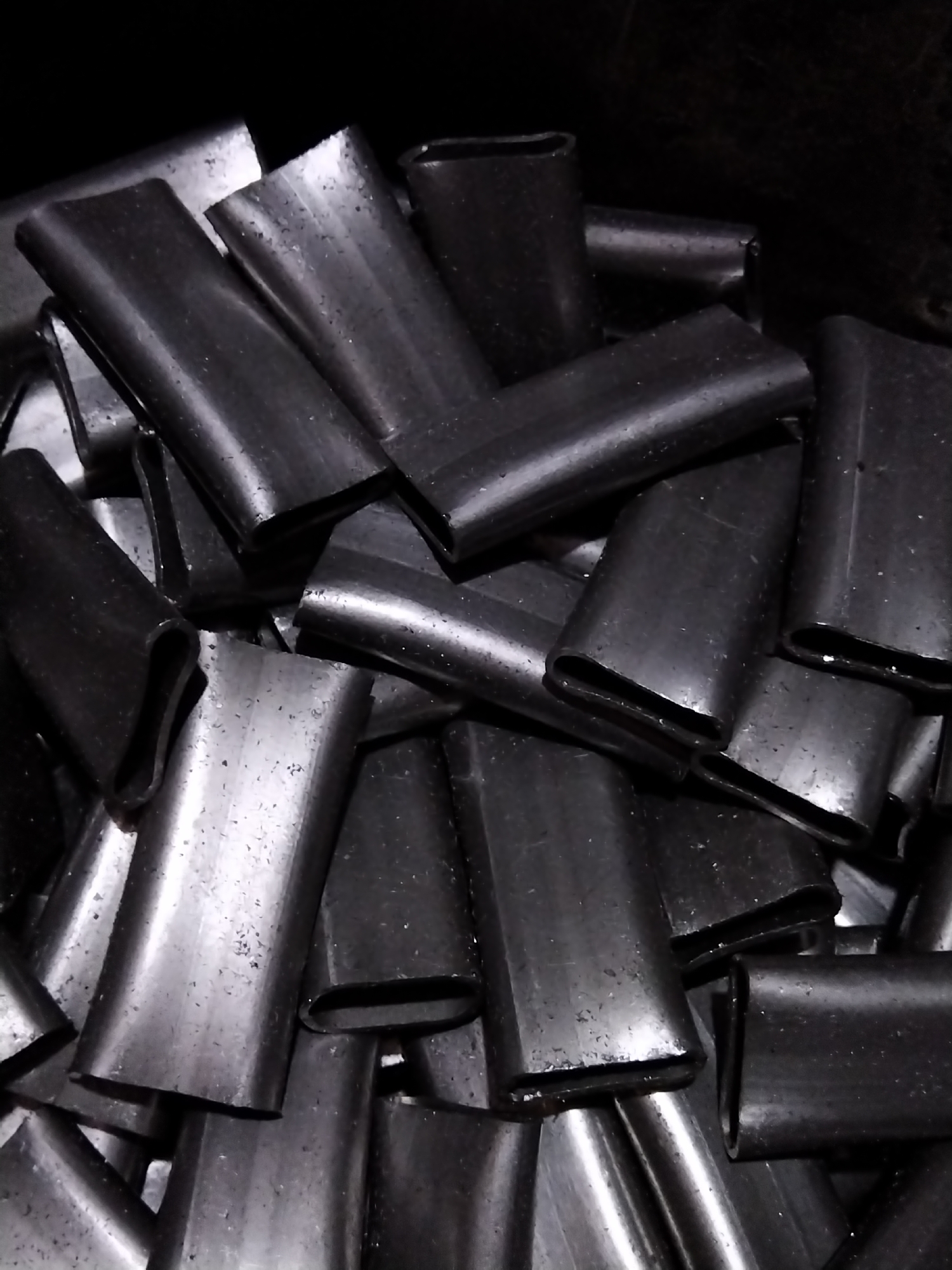 The image shows a pile of dark, cylindrical objects with flattened ends. These objects appear to be metal sleeves, possibly used for crimping or joining cables. They are arranged in a dense cluster, with some overlapping and obscuring others. The lighting is dim, creating a high-contrast scene with deep shadows and bright highlights reflecting off the metallic surfaces. The background is black, suggesting a dark, enclosed space. There is no indication of people, actions, or interactions. The image does not contain any contextual information that would allow for its placement in Pargolovo, Russia, or any other specific location.