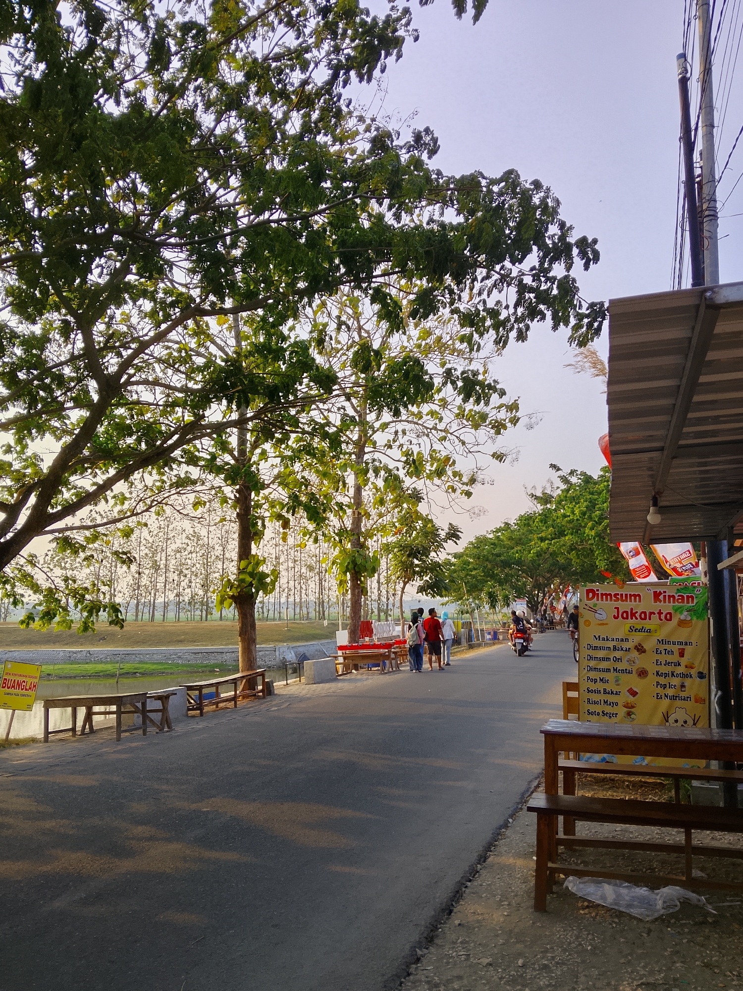 The image depicts a street scene in Ngadrin, Indonesia. In the foreground, a paved road runs along the right side of the frame, bordered by a sidewalk and a stall. The stall has a corrugated metal roof and displays a yellow banner advertising "Dimsum Kinan Jakarta," listing food and drink items. Several wooden tables and benches are set up in front of the stall.
To the left of the road, a body of water is visible, lined by a low concrete barrier and a grassy embankment. Further back, a line of trees creates a natural backdrop, with a dense, younger forest of trees visible beyond them. Large, mature trees with green foliage cast shadows on the road, indicating it is likely late afternoon or early morning.
Several individuals are present. A group of three people, two women and a man, are walking on the road, heading away from the camera. A motorcyclist is also seen riding down the street.
The sky is clear and light blue, with a few wispy clouds. The overall impression is of a quiet, somewhat rural street with food vendors and natural elements.