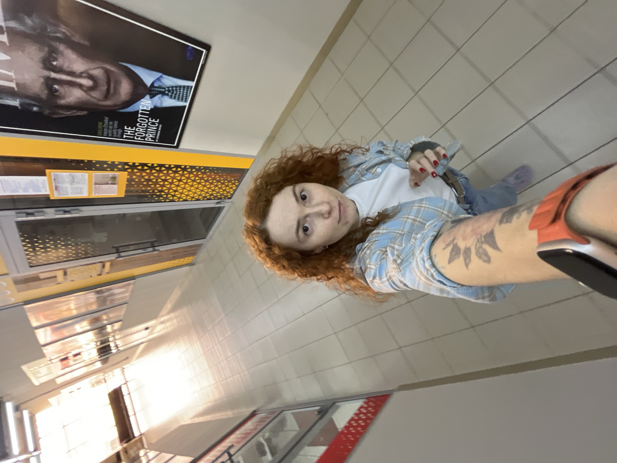 The image is a low-angle selfie-style shot of a young woman taken in an indoor public space, likely a shopping center or large building in Chaikovsky, Russia. The photo is oriented 90 degrees counter-clockwise, causing the tiled floor to appear as a wall.
The subject is a woman with curly red hair, looking directly at the camera with a neutral expression. She wears a light blue and white plaid button-up shirt over a white t-shirt, blue jeans, and partially visible purple shoes. Her fingernails are painted red, and she has a tattoo of a floral or sun-like design on her right forearm.
The background features a light-colored tiled floor (appearing as a wall), a glass display case or storefront reflecting bright lights, and a decorative panel with a golden geometric pattern on a yellow surface. A prominent large poster is visible on a dark background, featuring an older man's face. Key text on the poster reads "THE FORGOTTEN PRINCE." Below this, smaller text includes phrases like "EXCLUSIVE," "PRINCE CHARLES," and "WORLD IN HIS OWN WORDS," indicating the poster is an advertisement, likely for a magazine, documentary, or book about Prince Charles. The lighting is predominantly artificial indoor light, with some bright reflections suggesting natural light from distant windows.