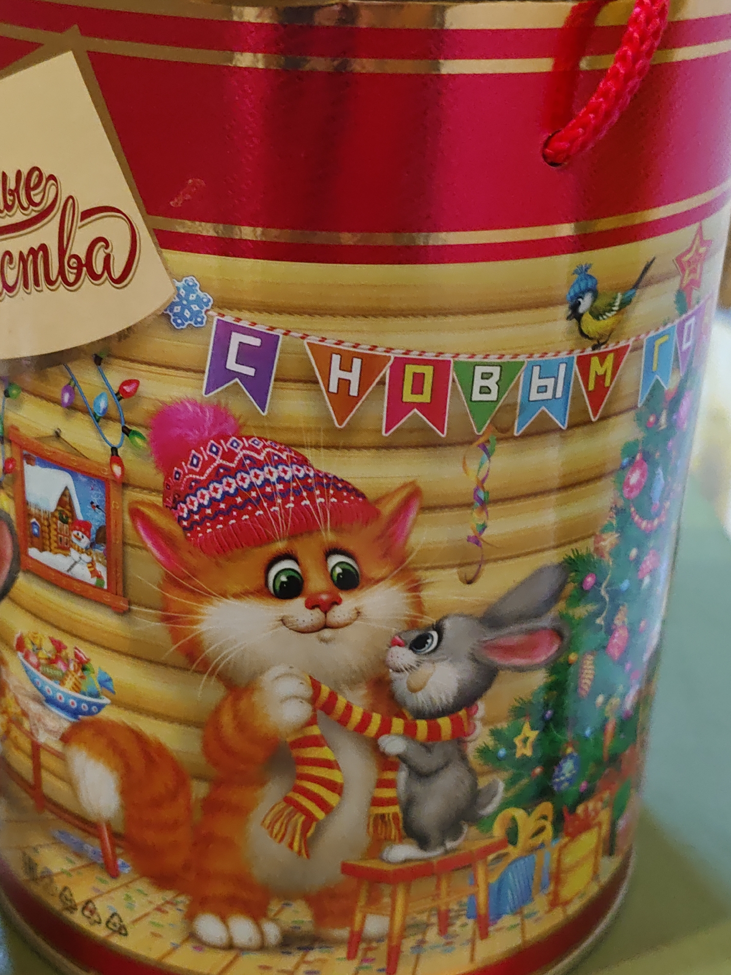 This is a close-up, eye-level, medium shot of a cylindrical container, likely a gift tin or cookie box, decorated with a festive Christmas theme. The background is a pale yellow, mimicking wood grain. A prominent red banner with gold stripes runs horizontally across the top. A bright red braided rope handle is attached to the top rim.
The main subjects are an anthropomorphic orange cat wearing a red and white patterned knitted hat and a grey rabbit. The cat is on the left, with large green eyes, a pink nose, and a fluffy white chest. It is wearing a red and yellow striped scarf wrapped around its neck, with the ends draped over its chest. The rabbit is to the right of the cat, facing left, with large ears and eyes. It appears to be interacting with the cat, possibly helping it adjust its scarf.
Below the animals, a wooden table or shelf is depicted, with a small red stool to the right. To the left of the cat, a framed picture of a snowy scene with a gingerbread house and a snowman is hanging on the wall. Colorful Christmas lights are strung above the picture.
Further to the right, a decorated Christmas tree is visible, adorned with colorful ornaments and a yellow star on top. Presents are stacked at the base of the tree.
Across the mid-section of the container, a banner of small triangular flags spells out "С НОВЫМ ГОДОМ" (Happy New Year in Russian). A small blue bird wearing a knitted hat is perched on the banner to the right of the text. A snowflake decoration is positioned above the cat's head.
The overall scene is brightly colored and whimsical, evoking a sense of holiday cheer. The image does not depict any recognizable city or location.