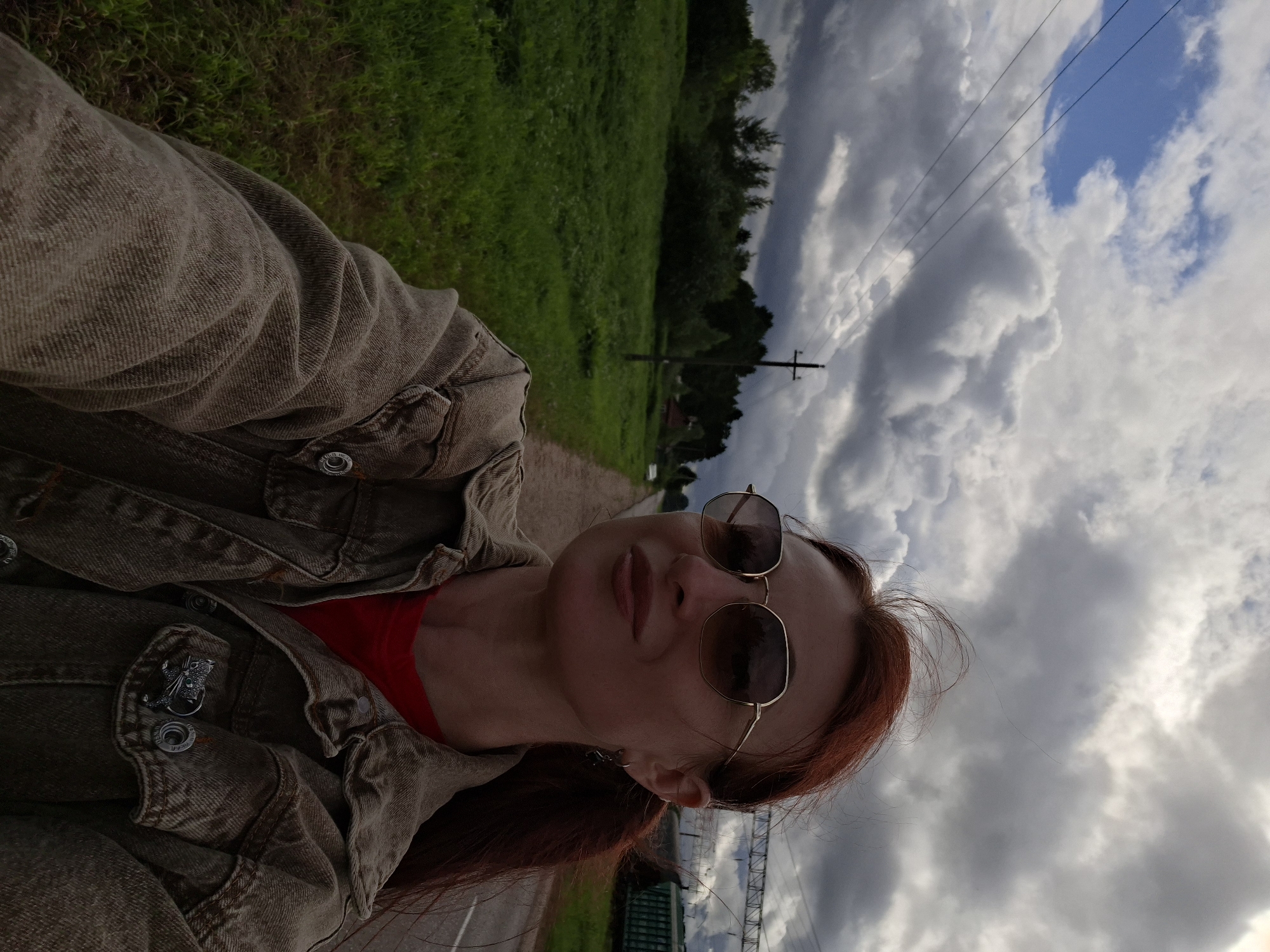 A selfie is taken by a woman with red hair and sunglasses, wearing a denim jacket and a red shirt, standing on an asphalt road. She is outdoors, with a field of green grass on her left and trees in the background. Power lines are visible above, and a train car can be seen in the far right of the image. The sky is cloudy, with a mix of dark and light clouds. The image does not provide enough context to determine the specific location within the city of Smaliavichy, Belarus.