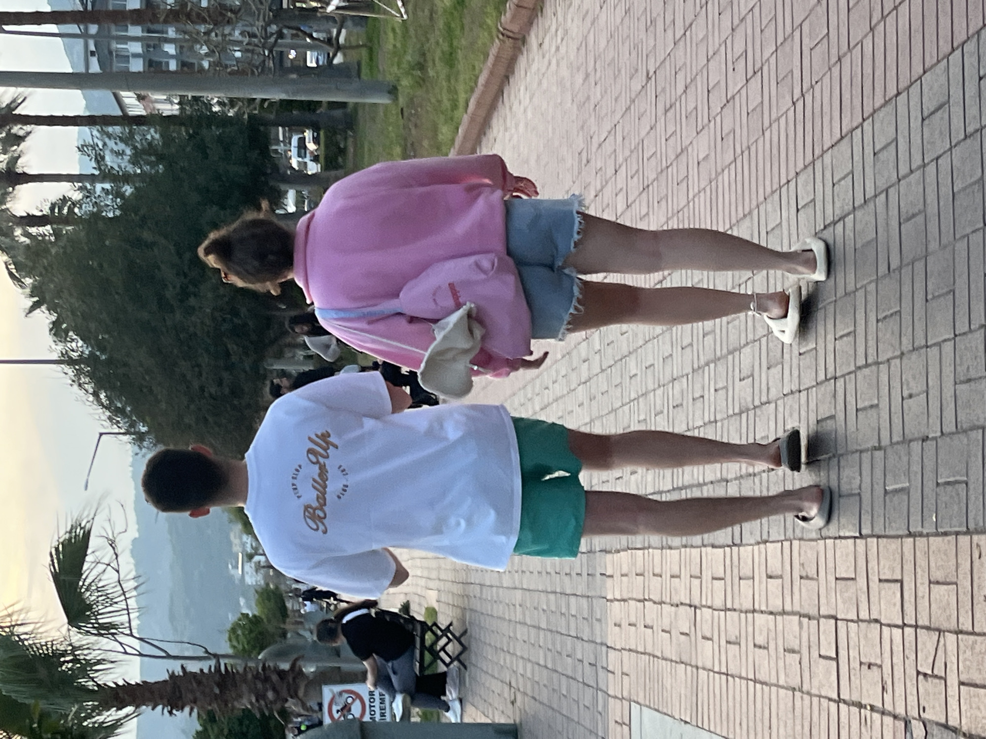 Two individuals are walking side-by-side on a brick sidewalk. The first person, presumably female, wears a pink jacket and jean shorts, with white sandals. The second person, presumed male, wears a white t-shirt with the text "Baller Up" written on it, green shorts, and grey sandals.
The location appears to be Alanya, Turkey, based on the visible landscape features. A palm tree is partially visible on the left side. In the distance, there are hills and a body of water, most likely the Mediterranean Sea. Buildings are visible in the mid-ground. People and street furniture can be seen in the surrounding area.