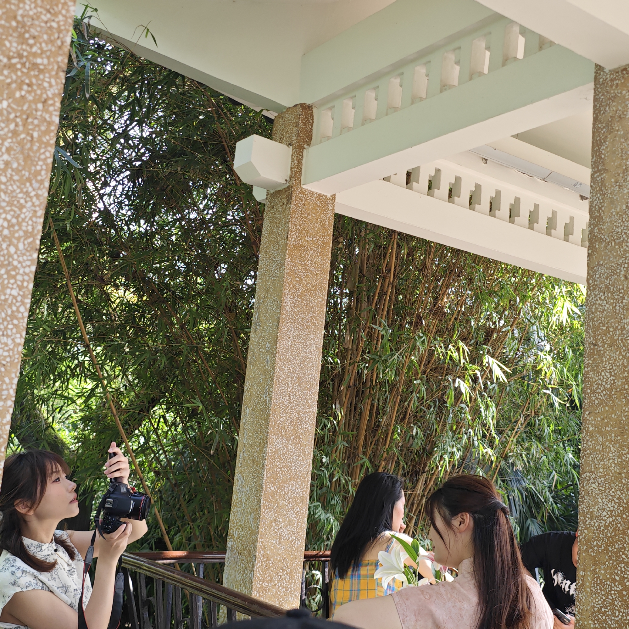 The image shows a group of people, likely at a photography event, in Yuzhong District, China. A woman on the left is holding a camera and appears to be taking a picture. Two other women are seated near a railing, with one holding white flowers. The scene is framed by beige stone pillars supporting a roof, and behind the group is a backdrop of dense bamboo foliage.
