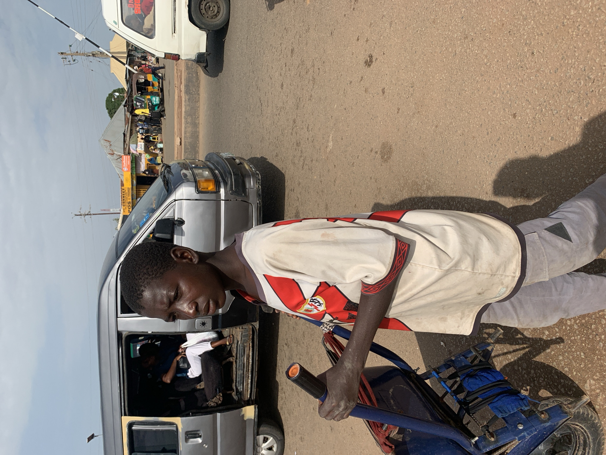 The image depicts a bustling street scene in Gura Top, Nigeria, likely during daytime given the strong shadows cast on the asphalt road. The foreground is dominated by a young Black male, appearing to be in his late teens or early twenties, who is leaning forward and pushing a blue, three-wheeled cart. He is wearing a light-colored t-shirt with red and black trim and grey trousers. His expression is one of concentration or weariness.
Behind him, a silver minivan is parked, with its side door open. Inside the minivan, several other individuals are visible, including a person in a white shirt and dark trousers seated in the passenger area. Further down the road, other vehicles are present, including a white van and a yellow auto-rickshaw, suggesting a busy traffic environment. Numerous people are seen standing around or walking, possibly shoppers or commuters, in front of roadside stalls or shops with corrugated metal roofs.
The overall atmosphere appears to be that of a typical market day or transportation hub. The ground is asphalt, showing signs of wear and tear, with some debris scattered on it. The sky is visible in the upper portion of the image and appears to be clear. No specific text is clearly legible in the image, though there are some signs on the roadside structures which are too small to be identified. The scene captures a moment of everyday life and commerce in a Nigerian town.