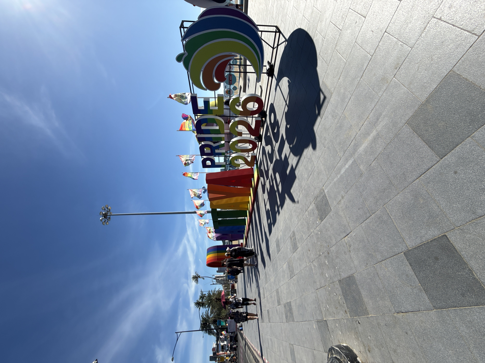 The image shows a large, colorful "PRIDE 2026" sign on a sunny day in Pattaya City, Thailand. The sign is composed of large, individual letters painted in rainbow colors, with "PRIDE" displayed prominently and "2026" below it. To the right of the sign is a large, stylized rainbow wave graphic. Several rainbow and pride flags are visible behind the sign, fluttering in the breeze. The sign is situated on a paved walkway, with people walking in the background. In the distance, palm trees and buildings are visible, suggesting an urban coastal environment. The sky is a clear blue with some wispy clouds. The overall scene conveys a festive atmosphere related to Pride events.