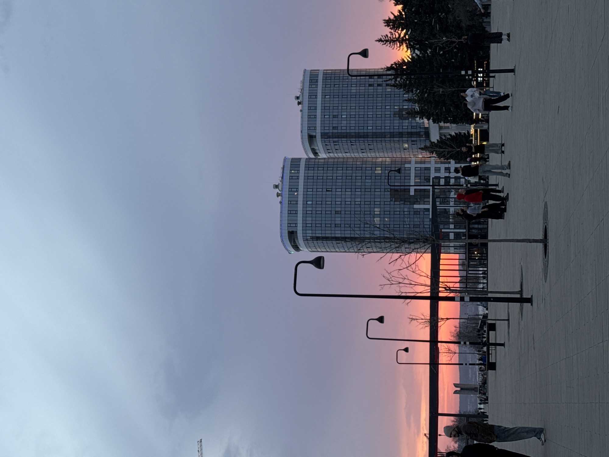 The image shows a cityscape during a sunset. Two high-rise, curved buildings with glass facades dominate the view. In front of the buildings is a paved plaza where several people are walking and standing. The sky transitions from a pale blue-gray to shades of pink and orange near the horizon. Street lights line the plaza. The location is Izhevsk, Russia.
