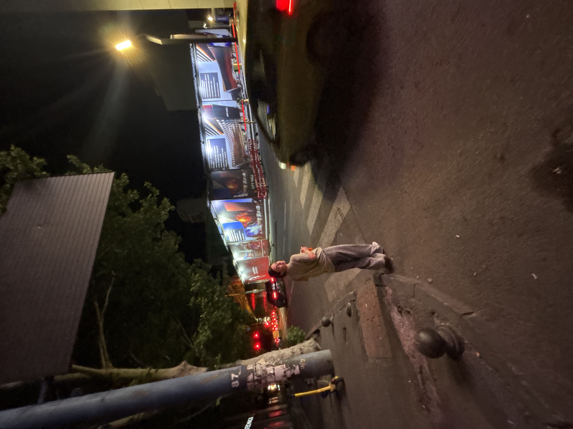 The image depicts a nighttime scene on a city street, likely in Tbilisi, Georgia, given the visible signage. A young woman with dark hair, wearing a light-colored t-shirt and grey sweatpants, stands on a pedestrian crossing, looking over her shoulder towards the camera. She holds a drink in her right hand. In the background, construction fencing with large advertisements is visible, along with streetlights and traffic lights. A yellow scooter is parked on the sidewalk to the left, next to a utility pole with markings including "87". Cars are present on the road, and a yellow car is partially visible in the foreground, appearing to be in motion. The overall lighting suggests it is dark, with artificial light sources illuminating the scene.
