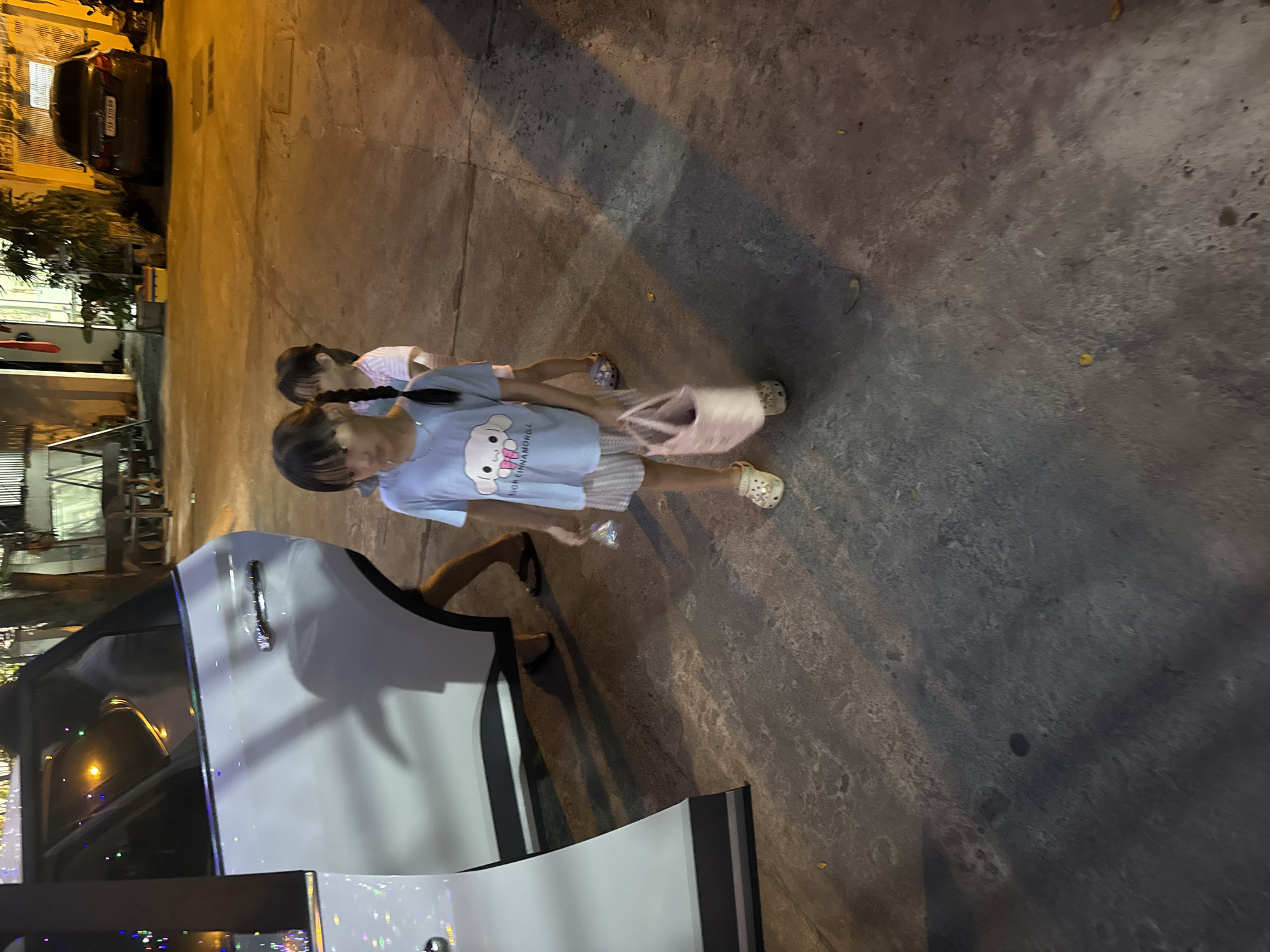 Two young girls stand beside a white car in Nam Nha Trang, Vietnam. One girl, closer to the car, wears a light blue shirt with a cartoon character and striped shorts, carrying a pink bag. The other girl is slightly behind, wearing a similar outfit. The car door is open, and the scene is illuminated by streetlights. A building and another car are visible in the background. The ground appears to be pavement.