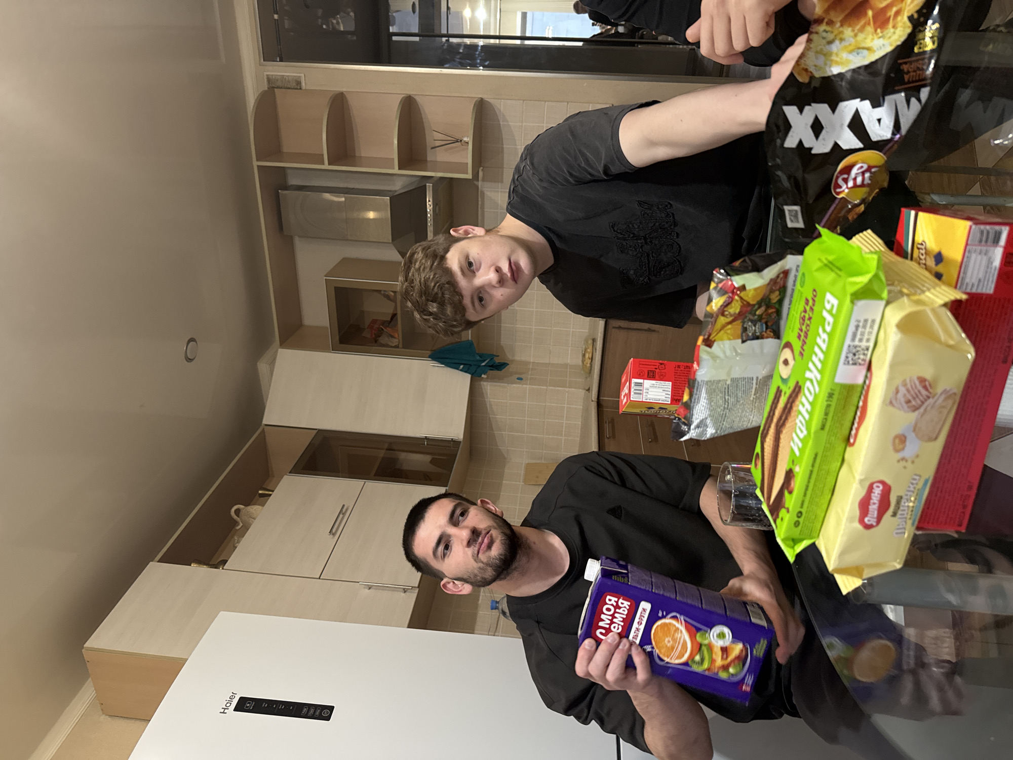 Two young men are seated at a glass table in a kitchen. One holds a juice box labeled "Moia Semia Multi-Fruit." Various snacks are on the table, including "Bryanskonfi" wafers, "Yashkino" cookies, and "Lay's Maxx" chips. The kitchen features light-colored cabinets and a white refrigerator with "Haier" visible on it. The photo was likely taken in Kazan, Russia, based on the product labels.