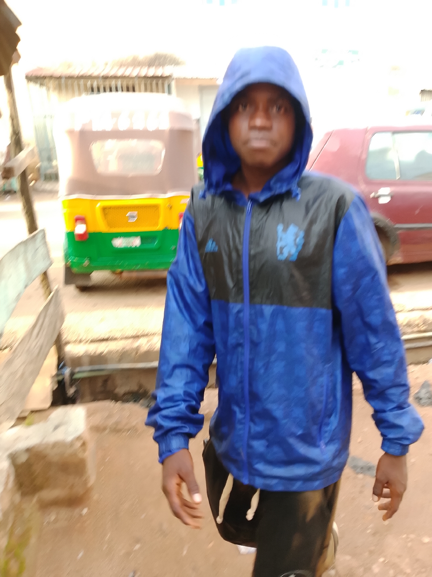 A young Black male, wearing a blue hooded jacket with a black panel across the chest featuring a blue logo, stands outdoors. He is also wearing black pants with white drawstrings. In the background, a yellow and green auto-rickshaw is parked, and a red vehicle is partially visible. The ground is dirt or gravel. The image appears to have been taken in Jos, Nigeria, judging by the context of previous images from the same location.