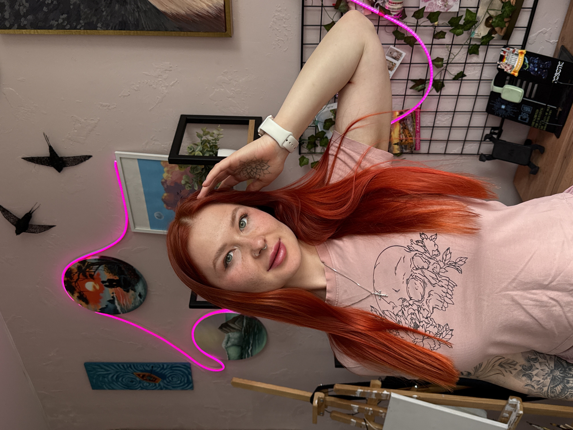 A woman with bright red hair is facing the camera. She has fair skin, freckles, and is wearing a light pink t-shirt with a skull design. She has a nose piercing and a silver necklace. Her left arm is raised, and she wears a white smartwatch on her wrist. A tattoo is visible on her left hand and arm.
The background shows a decorated interior wall with various art pieces and decorative items. There is a painting at the top left corner, and two black decorative bird figures are attached to the wall. Below the birds are three round art pieces and a rectangular blue art piece. A pink neon light is shaped around these art pieces. There is a black-framed picture of plants behind the woman's head and a wire grid with ivy vines and decorative items. A black box is on a wooden surface near the wire grid. An easel is visible at the bottom of the frame.
The image does not contain any information to determine the city of Rostov-on-Don, Russia.