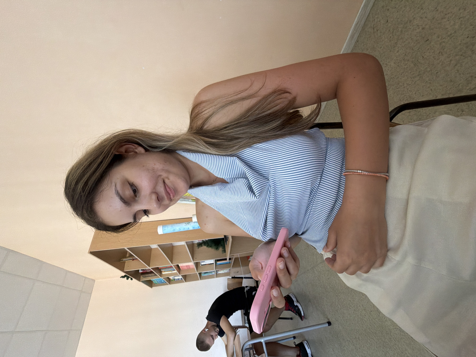 In a brightly lit classroom with light peach walls and a suspended ceiling, a young woman with long, light brown hair sits in a black chair, holding a pink smartphone. She is wearing a blue and white striped halter top and a light-colored skirt. Her gaze is directed downwards towards the phone. To her left, a young man with short hair and wearing a black t-shirt and shorts is seated at a desk, looking down and wearing white earbuds. In the background, a bookshelf filled with books stands against the wall. The floor is tiled with a speckled pattern. The image appears to have been taken in Burgas, Bulgaria.
