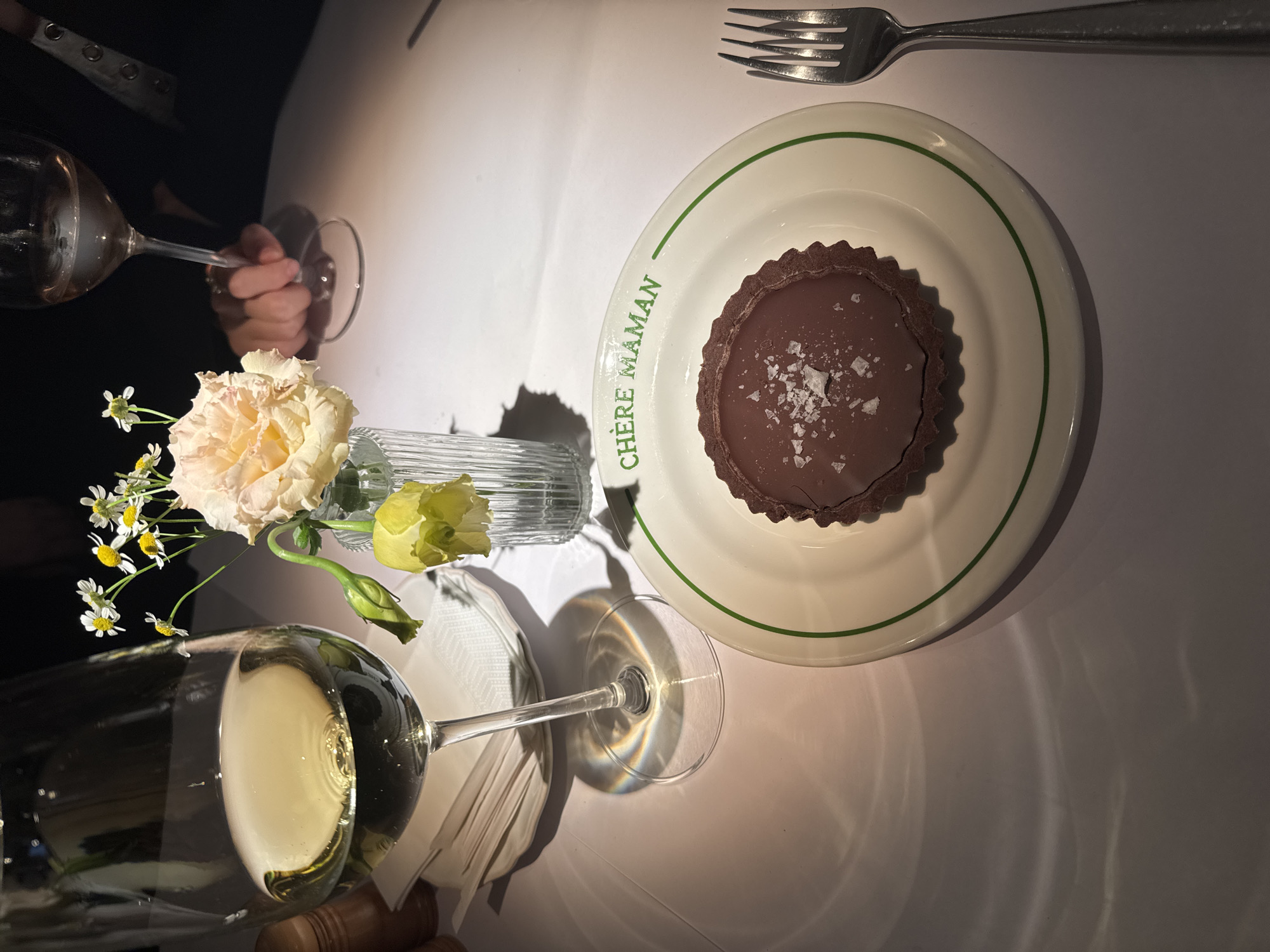 In an indoor setting, likely a restaurant, a table is set with a white tablecloth. The scene focuses on a dessert and a drink. In the foreground, a white plate with a green rim displays a chocolate tart topped with flaky sea salt. The plate bears the green text "CHÈRE MAMAN". To the left of the plate, a stemmed wine glass filled with a pale yellow liquid, likely white wine, is positioned. Behind the wine glass, a small bouquet of flowers in a clear glass vase includes a light pink rose, small white daisies, and a drooping yellow bud. A fork rests to the right of the plate. In the background, a second wine glass is held by a person whose hand and arm are visible. The lighting suggests a dimly lit environment, with light reflecting off the wine and casting shadows on the tablecloth. The image captures a moment of dining, emphasizing the dessert and the accompanying wine. The location in Moscow, Russia, is not discernible from the visual information provided in the image.