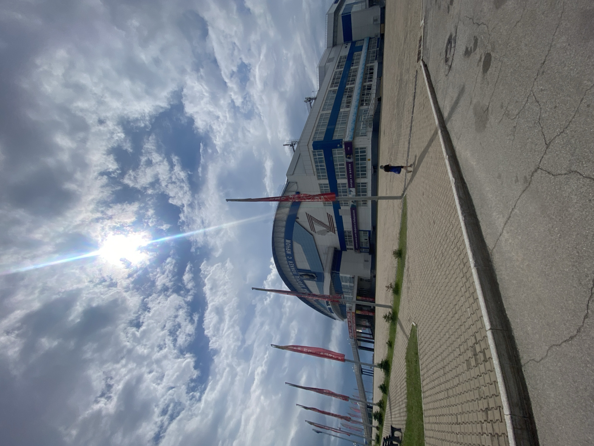 The image shows the "Platinum Arena" in Khabarovsk, Russia, a large multi-purpose indoor arena. The sky is cloudy with the sun shining through, creating lens flare. The arena has a distinctive curved roof and is white and blue with the letter "Z" prominently displayed on its facade. In front of the arena, there is a paved walkway with several red flags on poles. A person is walking on the walkway towards the arena. The ground in the foreground is asphalt with some cracks and puddles. The scene appears to be a public area outside the arena on a cloudy day.