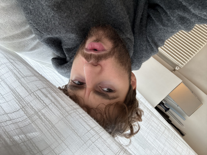 The image captures a close-up view of a young adult male, appearing to be in his 20s or 30s, with light brown, curly hair and a beard. His expression conveys mild confusion or inquiry, characterized by furrowed brows and slightly parted lips. He is wearing a dark grey hooded garment. A light-colored, textured fabric, possibly a curtain or blanket, partially obscures the upper right portion of the frame and drapes over his head. The background on the left reveals a white wall with a horizontal, multi-finned radiator. Above the radiator, a white shelf displays a rectangular reflective object, a black-bristled drum brush, and several other elongated tools. The architectural features, specifically the radiator design, are consistent with an indoor domestic setting, likely an apartment, in Paris, France.