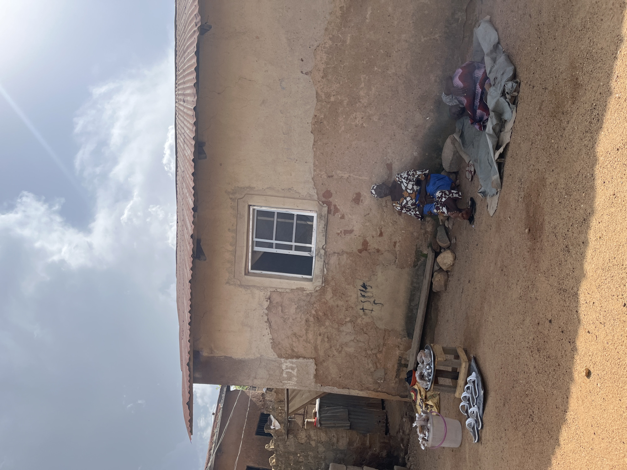 In this outdoor scene, possibly in Jos, Nigeria, two individuals are visible near a simple, aged building with a corrugated metal roof. The building's walls are made of rough, textured plaster, showing signs of wear and discoloration. A single window with a white frame is set into the wall.
One person, dressed in a patterned fabric and blue garment, is seated on the ground, partially covered by a gray cloth. Another person, also in patterned clothing, is lying down next to them, partially obscured by the same cloth and some rocks. A pair of flip-flops lies near the seated individual.
In the foreground, on a wooden stand, are various items for sale, including what appear to be small plates of food or goods, and several pairs of sandals. A pale pink bucket sits nearby. The ground is sandy and dry.
The sky above is bright, with scattered white clouds, suggesting daytime. The sunlight casts shadows, indicating it is likely morning or afternoon. The overall impression is of a humble, possibly market-like setting on a clear day. No visible text is present in the image.