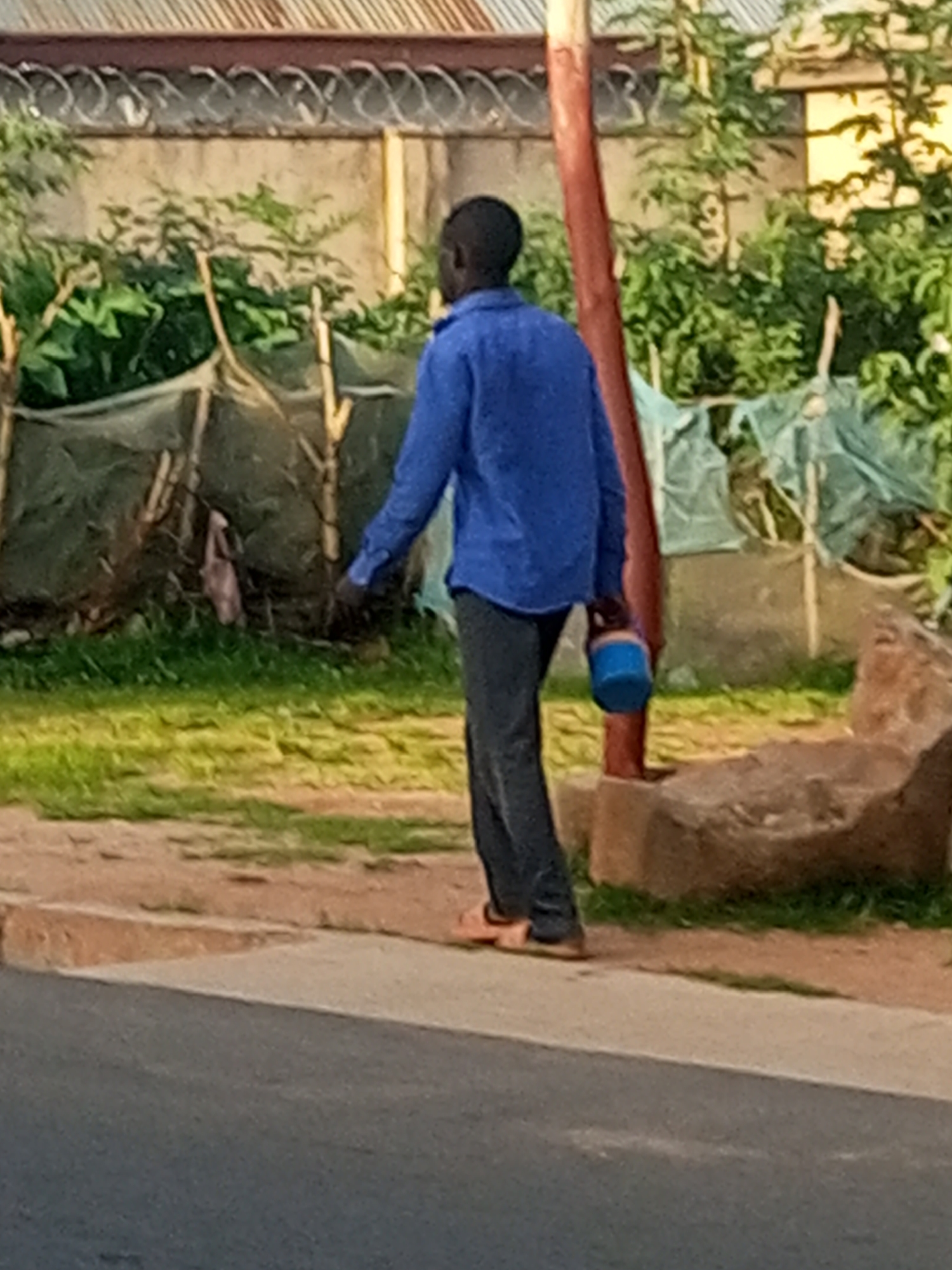 A person wearing a blue long-sleeved shirt and dark pants walks along a dirt path next to a paved road. They are carrying a blue bucket in their right hand. Behind them, there is a fence with barbed wire and some greenery. A large rock is visible to the right of the person. The scene appears to be in an outdoor urban or semi-urban setting.