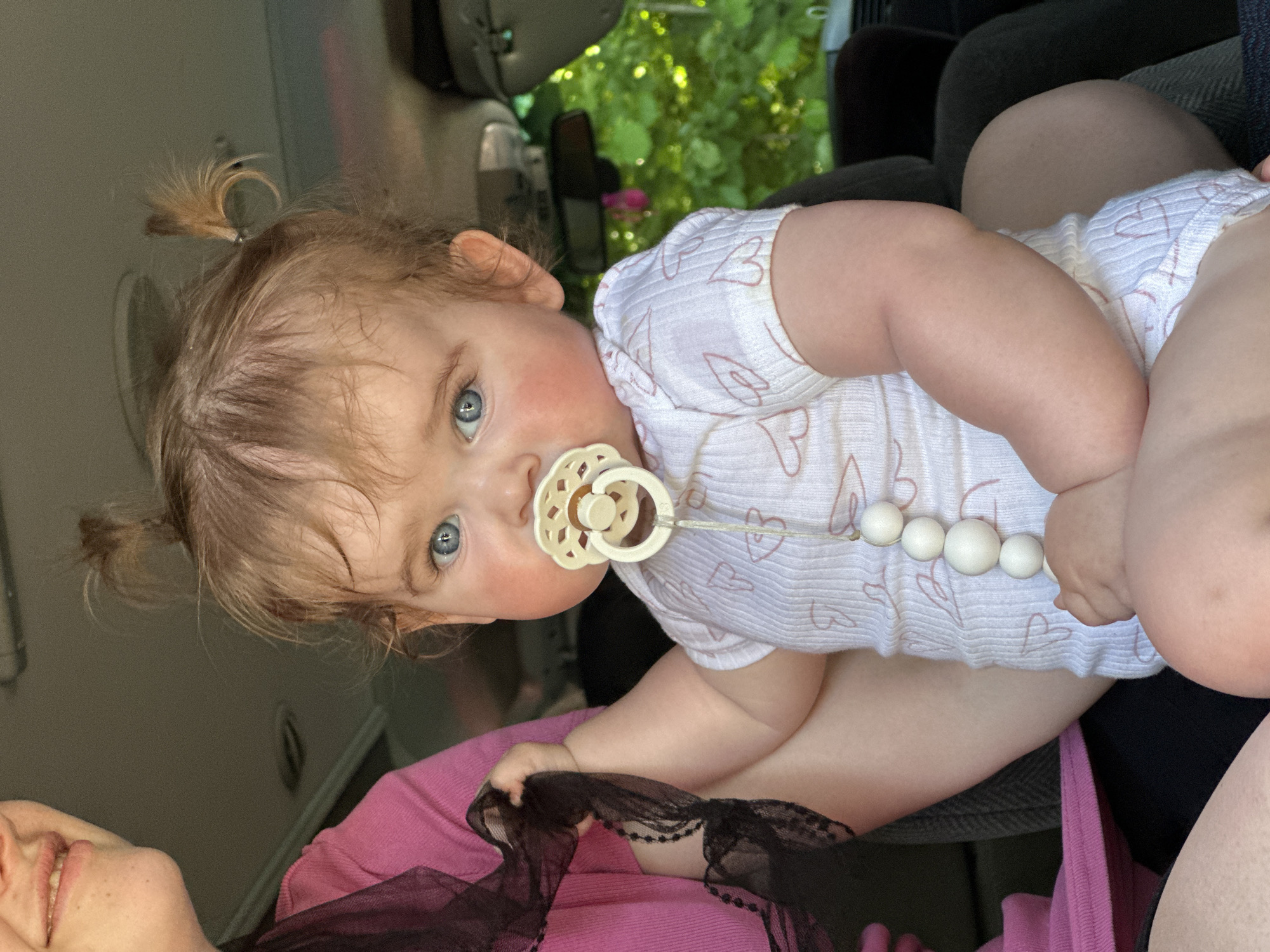A baby girl with blonde hair styled in pigtails sits on someone's lap in a vehicle, possibly a van. She has blue eyes, pink cheeks, and a pacifier with a white beaded chain. She wears a white onesie with heart patterns. The adult holding her wears a pink top and a black lace garment is visible in the foreground. The background shows the interior of the vehicle and glimpses of greenery outside the window. The scene appears to be indoors, possibly in a car traveling through Grünberg, Germany based on the greenery and architectural cues suggesting the region. The image composition features a slight tilt.