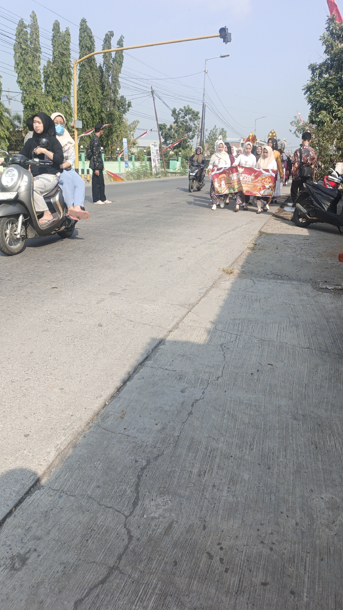 In Sumengko, Indonesia, on a sunny day, a group of people are participating in a parade or procession. Two women are seated on a scooter, one driving and the other as a passenger, both wearing hijabs. Behind them, several women in hijabs and traditional Indonesian attire are walking, some carrying a banner with Indonesian text and images. The banner appears to be celebratory, possibly related to Indonesian Independence Day given the red and white flags visible in the background. One person in the procession wears an elaborate headpiece. Men are also present, including one in a patterned shirt walking behind the banner carriers, and another standing near a traffic light. The scene takes place on a paved road with a sidewalk on the right, lined with trees and some buildings in the distance. Two motorcycles are parked on the right side of the road. The overall atmosphere suggests a community event or celebration.