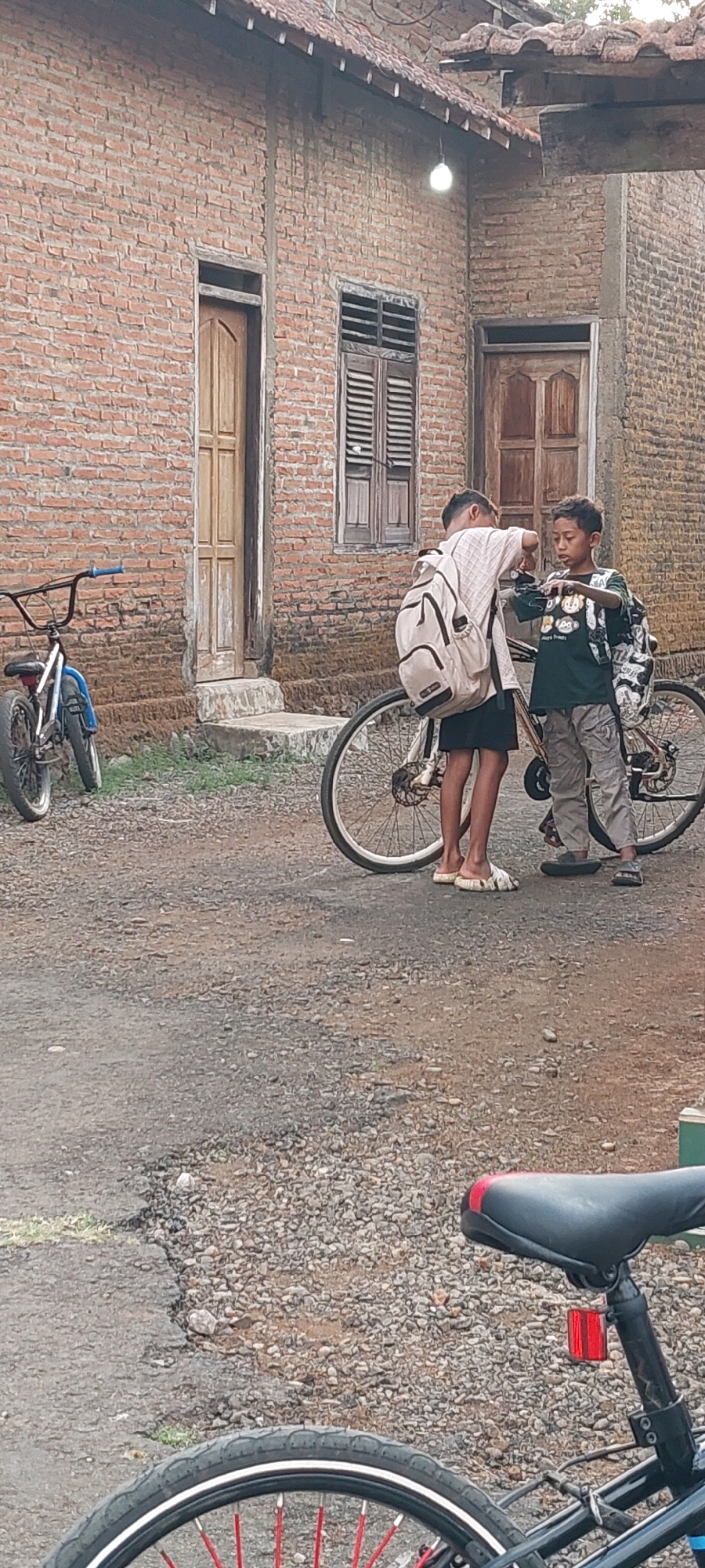 Two boys are standing next to a bicycle in front of a brick building in Keling, Indonesia. The building has a tiled roof, a wooden door, and a window with shutters. The ground is covered with gravel and dirt. The boy on the left is wearing a light-colored shirt and dark shorts, and has a backpack on. The boy on the right is wearing a dark t-shirt with a graphic print and light-colored pants, and also has a backpack. They appear to be interacting with each other. A second bicycle is visible to the left of the scene, and a third bicycle is partially visible in the foreground.