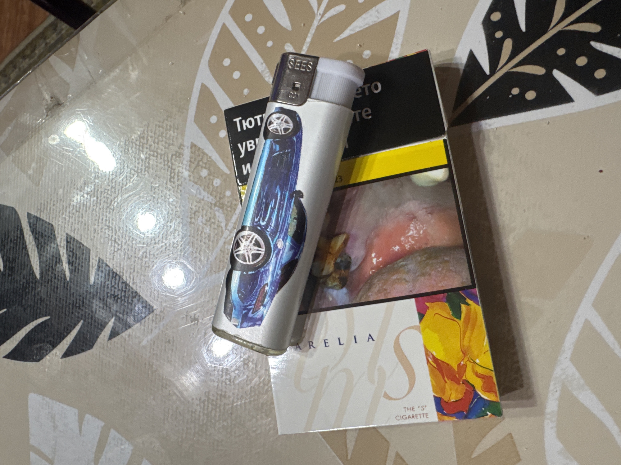 The scene is located in Sarnevo, Bulgaria. An overhead view captures two items resting on a patterned surface. A disposable butane lighter is positioned horizontally atop a cigarette pack. The lighter is silver-colored, featuring a graphic of a blue sports car. The metallic top of the lighter has "SEES" and "021" embossed on it, with a white igniter visible. The cigarette pack, partially obscured by the lighter, displays "SARELIA" and "THE 'S' CIGARETTE" on its front, which is predominantly white with large, stylized "S" curves and a colorful abstract design (yellow, orange, red, blue, green shapes) at the bottom. The visible side of the cigarette pack features a graphic health warning depicting an unhealthy mouth/gums, with Cyrillic text on a black background and a yellow stripe. The items rest on a light beige or off-white laminated surface adorned with a repeating pattern of stylized dark grey, beige, and white leaves or fronds. Multiple bright, circular reflections are present on the surface and objects, indicating an overhead light source. No individuals, actions, or interactions are depicted in the image.