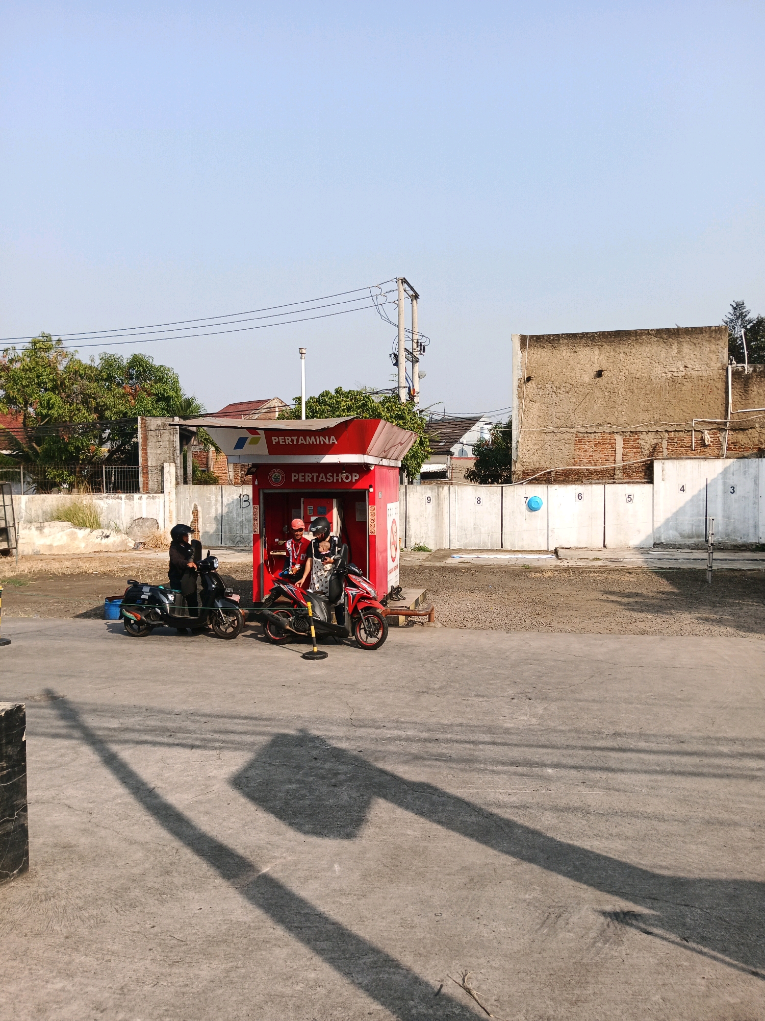 At a Pertamina gas station, two people on a black motorcycle and two people on a red motorcycle are interacting with a Pertamina attendant. The attendant, wearing a red shirt and a pink hat, is dispensing fuel to the red motorcycle. The black motorcycle is parked next to the red one. The scene is outdoors with a clear blue sky and sunlight casting shadows on the ground. The background shows a concrete wall with numbers from 3 to 9, and behind that, buildings and trees. The gas station itself is a small, red structure with "PERTAMINA" and "PERTASHOP" visible. The overall environment suggests a typical urban or suburban setting in Indonesia, possibly in Bandung City, Indonesia, given the presence of Pertamina, a state-owned oil and gas company prevalent in the country.